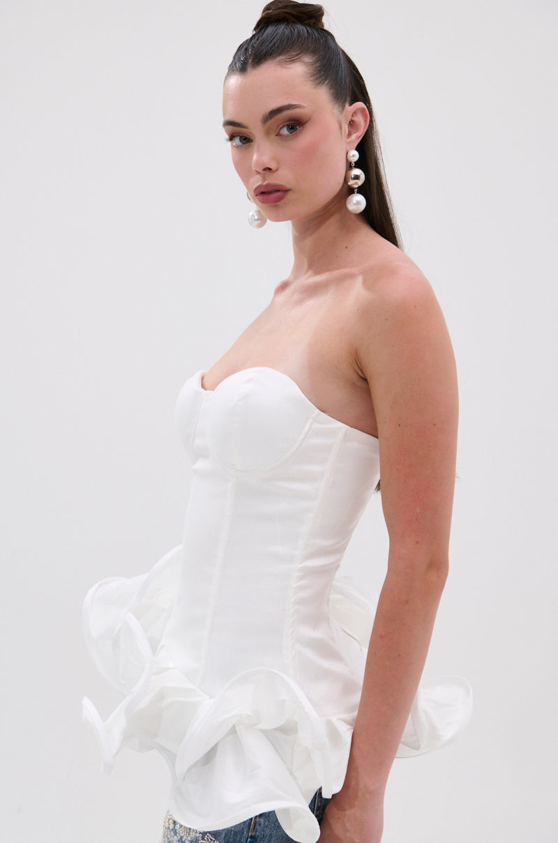 A woman with long brown hair in a high ponytail models the THE MORE YOU KNOW RUFFLE DETAIL CORSET TOP, a strapless sweetheart neckline piece with ruffle peplum, paired with large pearl earrings against a plain light background.