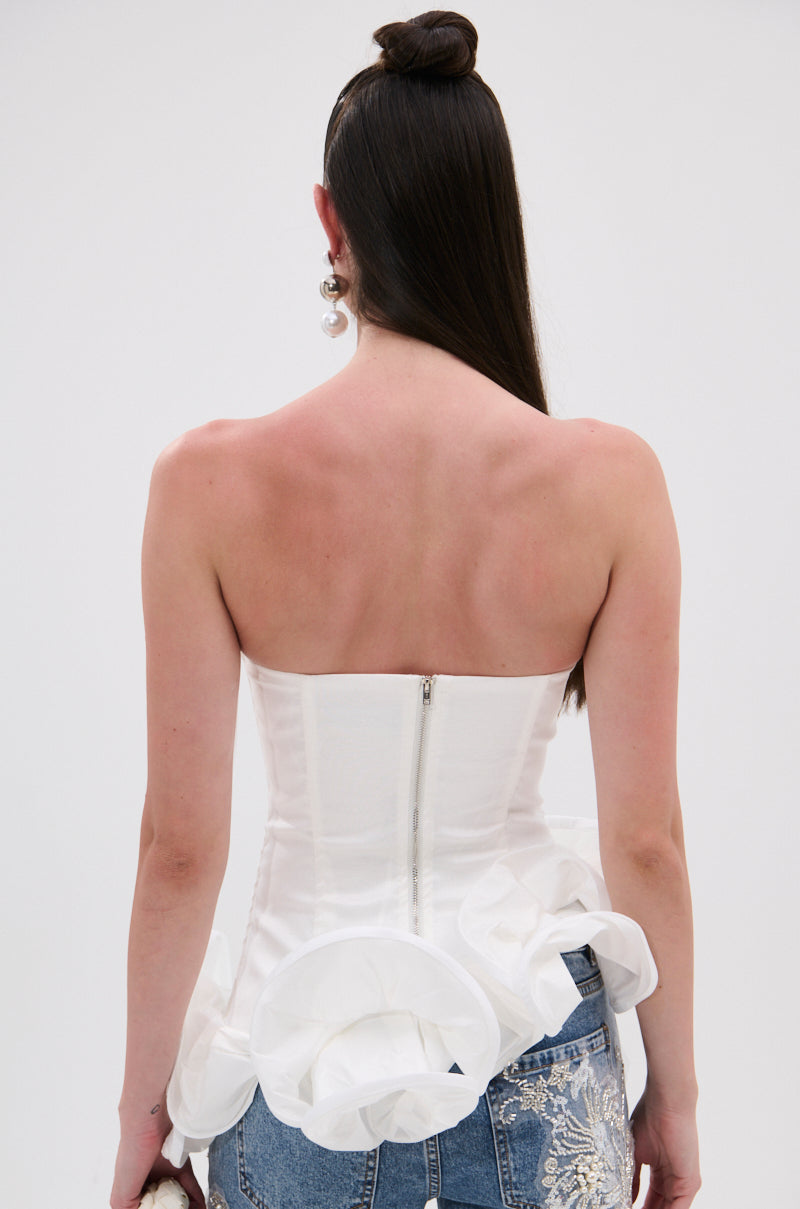 A person with long, straight dark hair faces away, wearing THE MORE YOU KNOW RUFFLE DETAIL CORSET TOP with a strapless sweetheart neckline and zipper, paired with blue jeans featuring white floral embroidery.