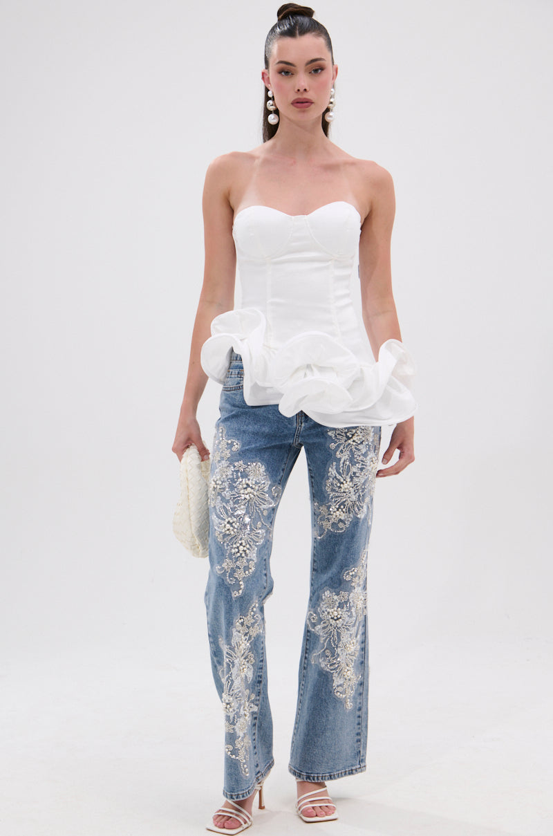 A woman stands against a plain white background wearing THE MORE YOU KNOW RUFFLE DETAIL CORSET TOP, blue jeans with white floral embroidery, white heeled sandals, statement earrings, and holding a textured white clutch.