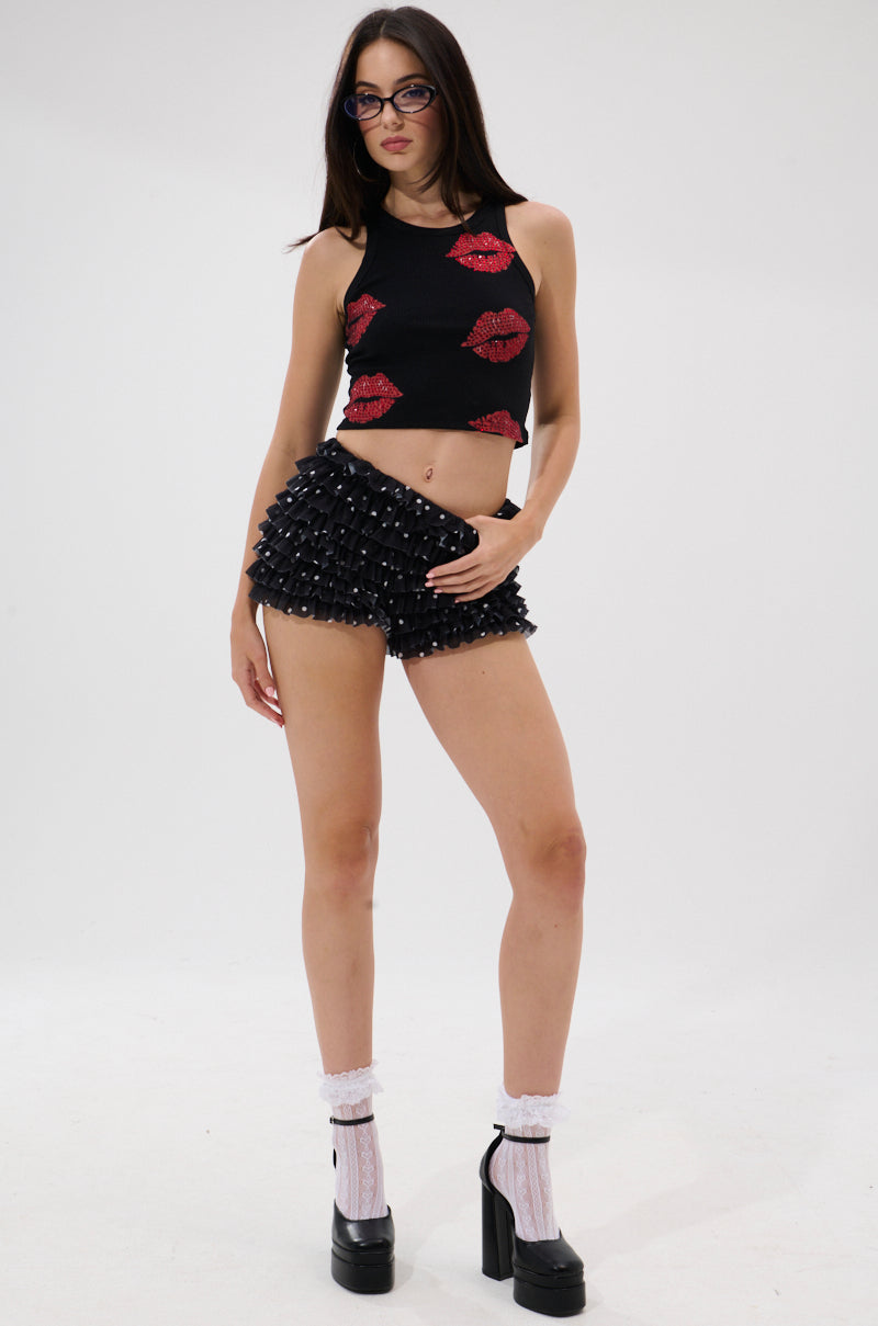 A woman in glasses wears the KISSY FACE TANK TOP IN BLACK with red lips, black textured high-waisted shorts, white frilly socks, and black platform heels while standing against a plain white background.