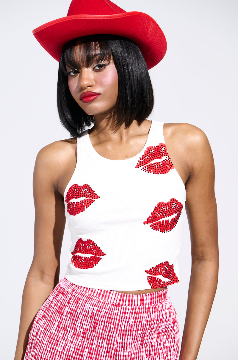 A woman sports a red cowboy hat and the AKIRA Label KISSY FACE TANK TOP, featuring a Y2K-inspired red sequin lips design. Her shoulder-length black hair and matching red lipstick enhance her vibrant ensemble, completed by a red and white checkered skirt against a plain white backdrop.