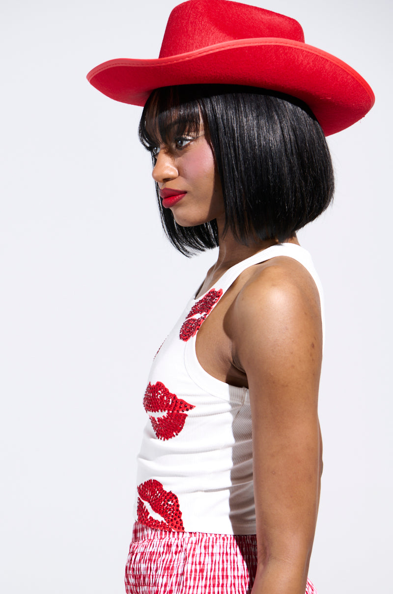 A person in a red cowboy hat, the KISSY FACE TANK TOP with red lip prints, and a red checkered skirt. They have shoulder-length black hair and wear red lipstick, standing against a plain white background.