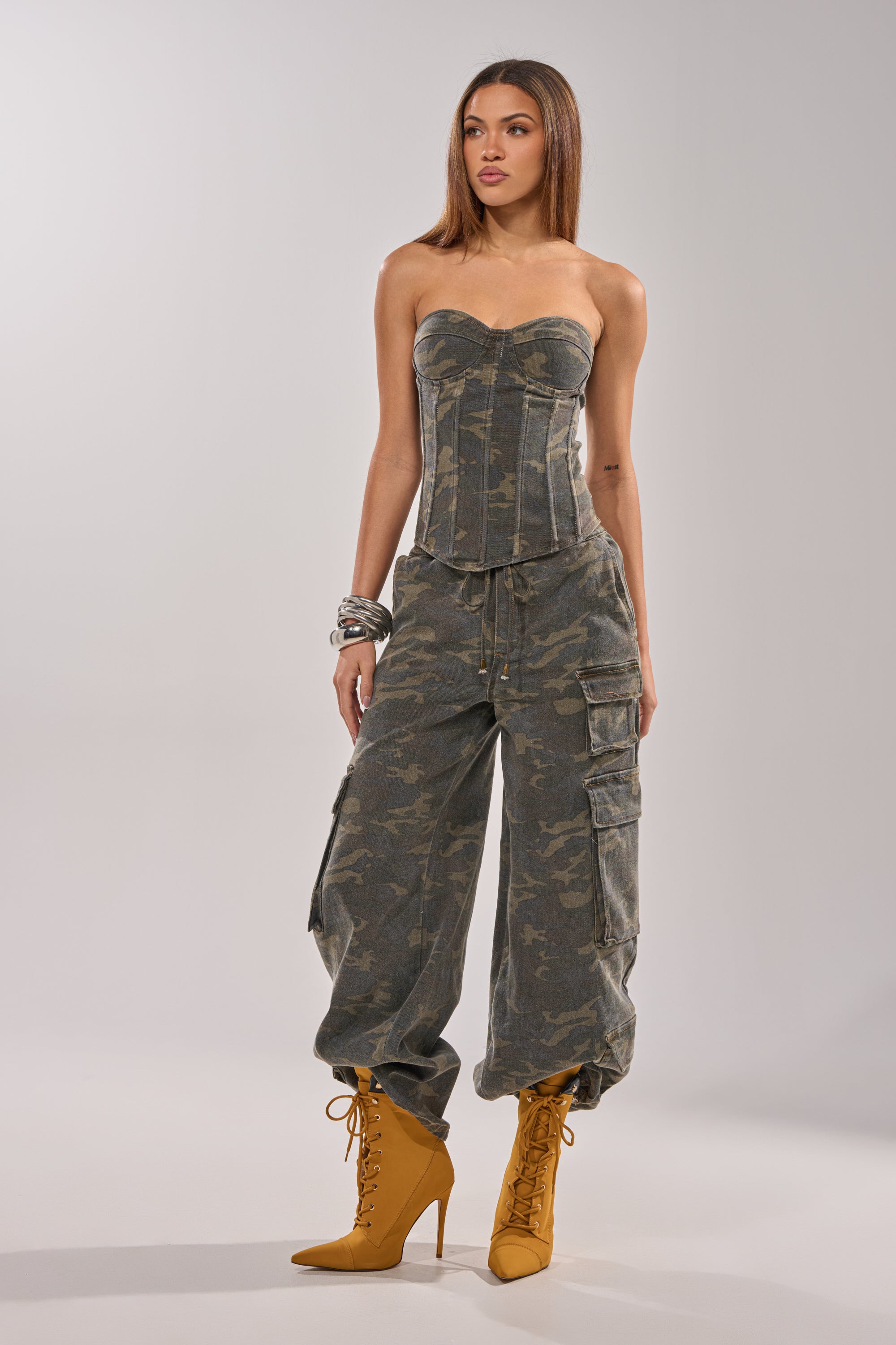 A woman poses against a light background in the DOWN AND DIRTY CAMO CORSET, featuring a strapless sweetheart neckline, paired with matching cargo pants, mustard yellow high-heeled boots, and bracelets. 6