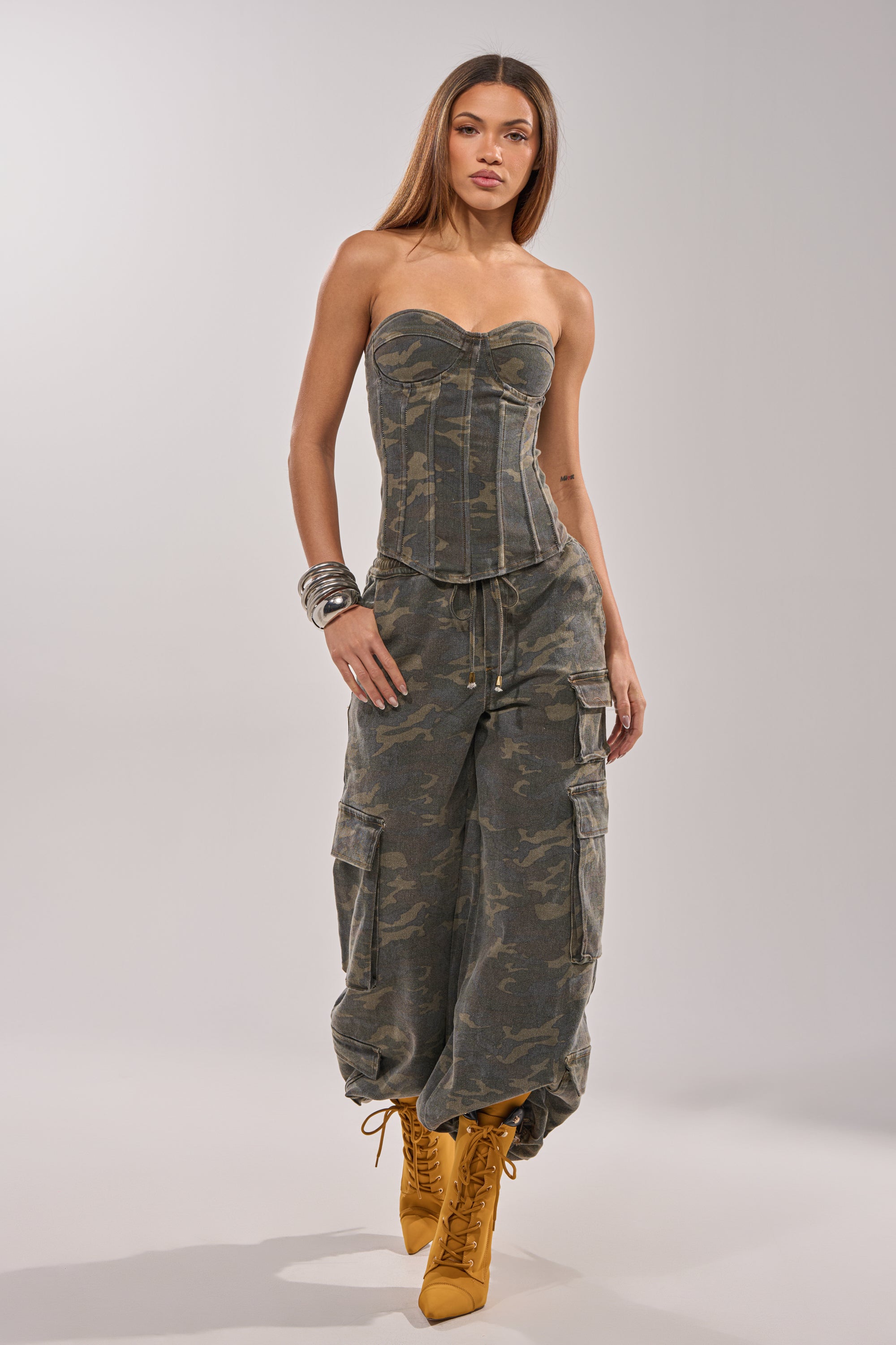 A woman wears the Down and Dirty Cargo Pant featuring a wide-leg camo print, styled with a strapless fitted camouflage corset top, yellow lace-up boots, and silver bangles against a plain light background. 0