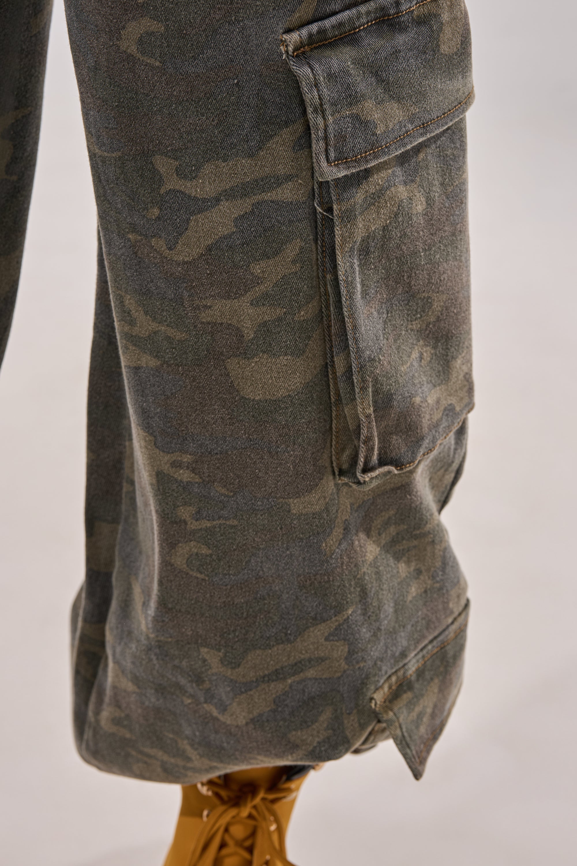 Close-up of the DOWN AND DIRTY CARGO PANT featuring a modern camo pattern, wide-leg silhouette, flap pocket, and visible stitching. The loose fit adds to its contemporary style, shown worn on a light surface. 9