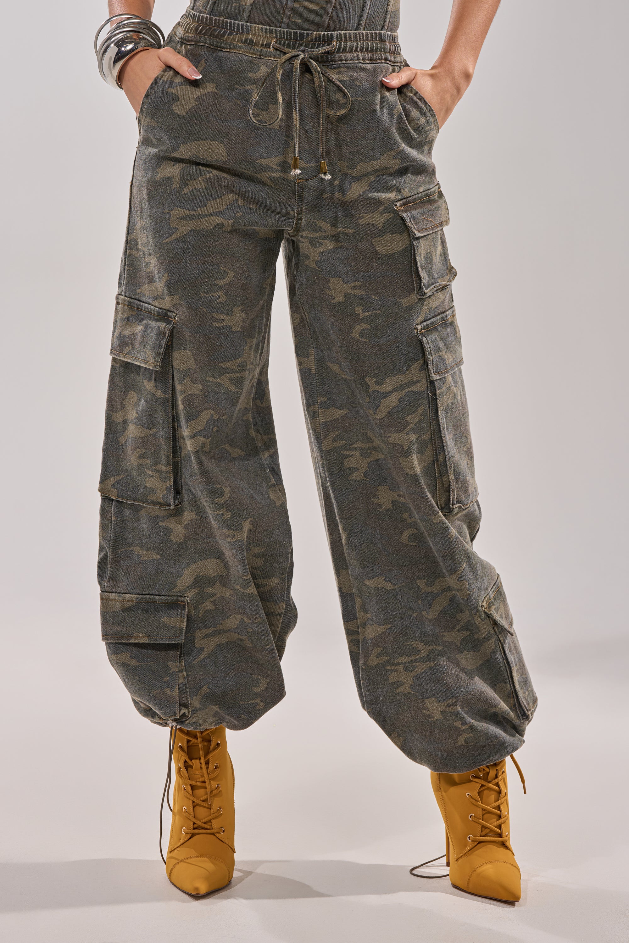 Wearing the DOWN AND DIRTY CARGO PANT—utility-inspired, camo wide-leg cargos with multiple pockets—a person pairs them with lace-up mustard yellow ankle boots, hands on hips, and metallic bracelets shining on one wrist. 1
