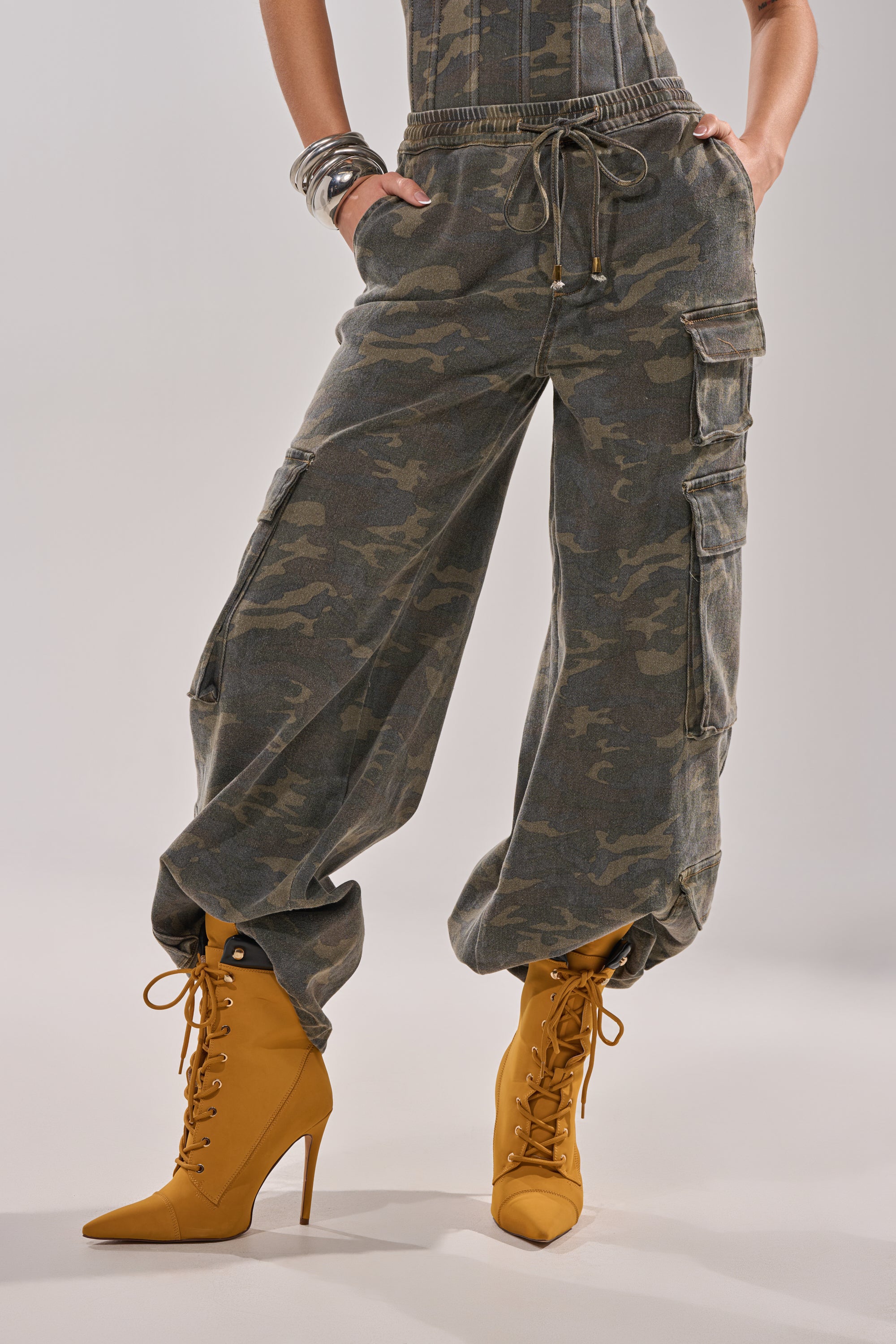 A person wears the DOWN AND DIRTY CARGO PANT, featuring wide legs, camo print, drawstrings, and pockets, paired with mustard yellow high-heeled lace-up boots. Their hands are in their pockets, and silver bangles decorate one wrist. 2
