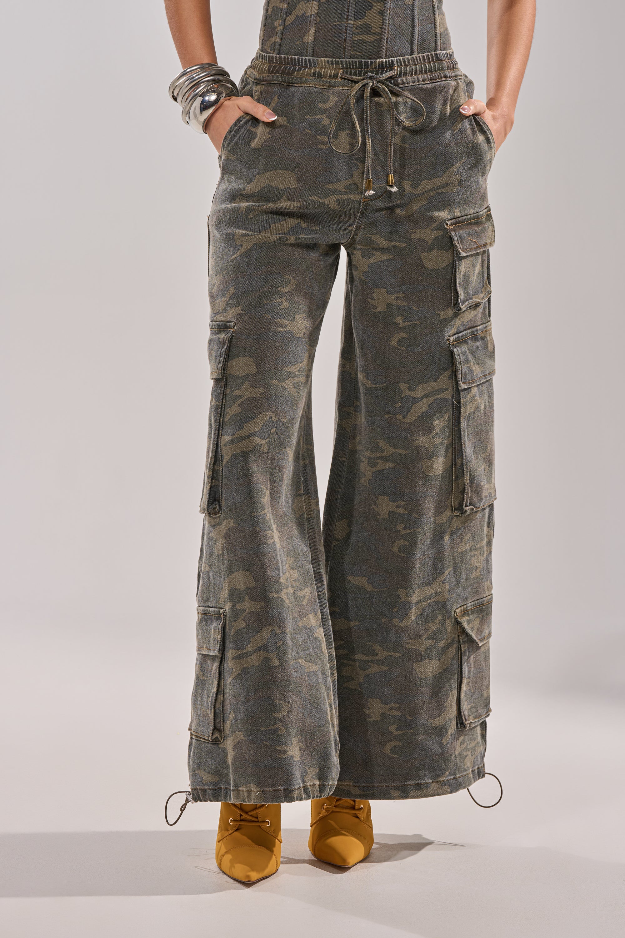 A person wearing the DOWN AND DIRTY CARGO PANT—utility-inspired camo with wide legs, large side pockets, and drawstrings at the waist and hem—paired with yellow pointed-toe heels and silver bangles. 3