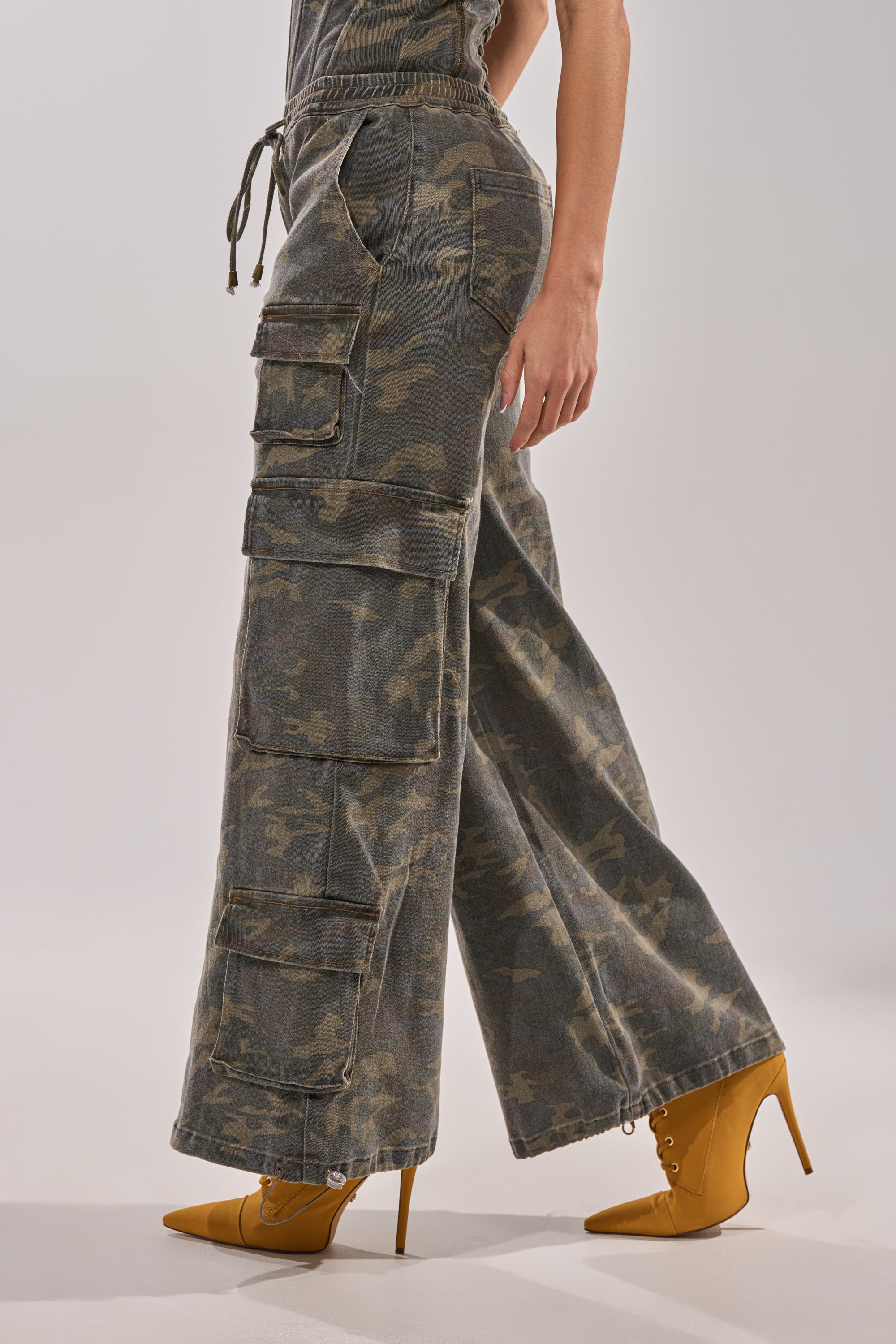 Someone models the DOWN AND DIRTY CARGO PANT, featuring a high-waisted, wide-leg camo look with large utility pockets, paired with yellow pointed-toe heeled boots against a light background. 4