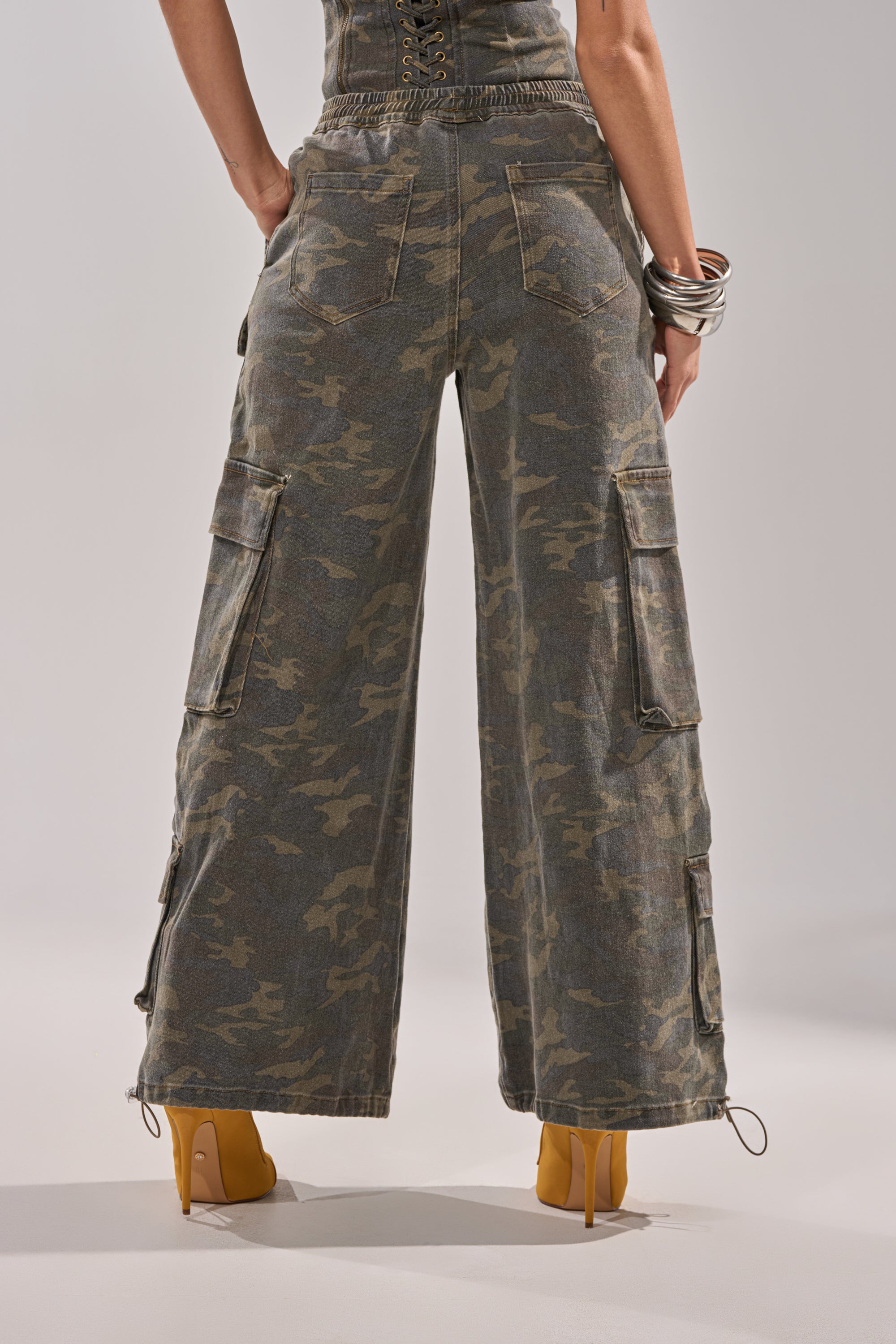 Wearing the DOWN AND DIRTY CARGO PANT, a person stands in yellow heels. The high-waisted, wide-leg camo pants feature side pockets and utility details. The image shows their back from waist to feet with arms partly visible. 5