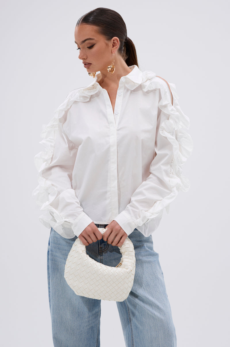 A woman wearing the ZIP ME OUT OF THIS BLOUSE with sculptural ruffles and light blue jeans holds a textured white handbag, her hair pulled back as she looks down against a neutral background. 0