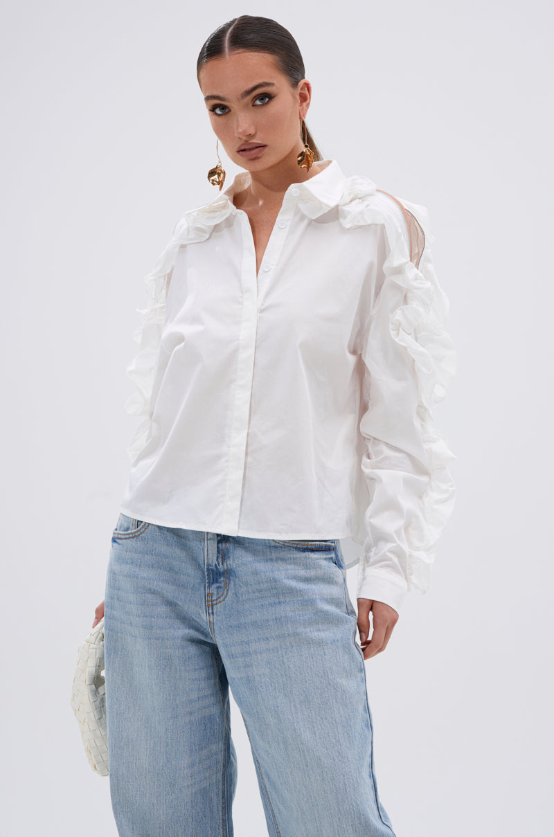 A woman wears the ZIP ME OUT OF THIS BLOUSE, a white button-up with sleeve ruffles, and light blue jeans as she stands confidently against a plain white background. 1