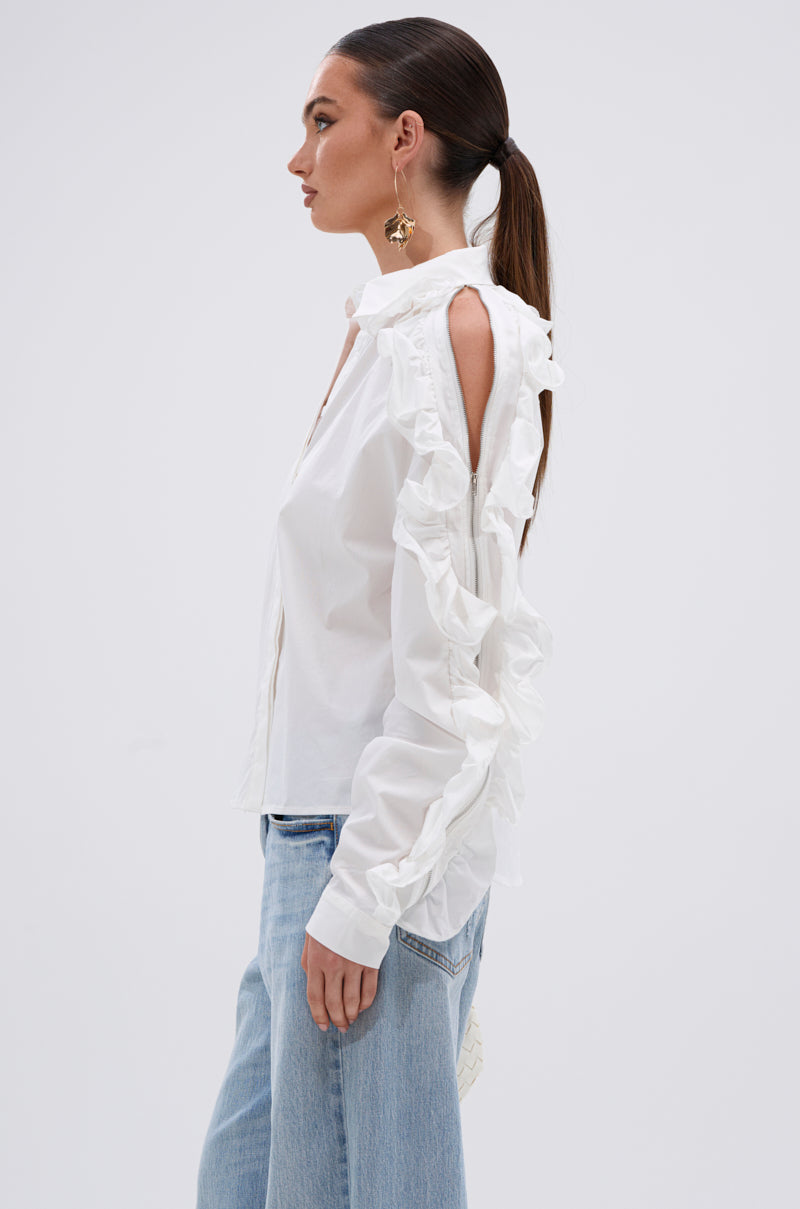A woman with a ponytail stands in profile against a plain white background, wearing the ZIP ME OUT OF THIS BLOUSE with ruffled open-shoulder sleeves, paired with light blue jeans. 2