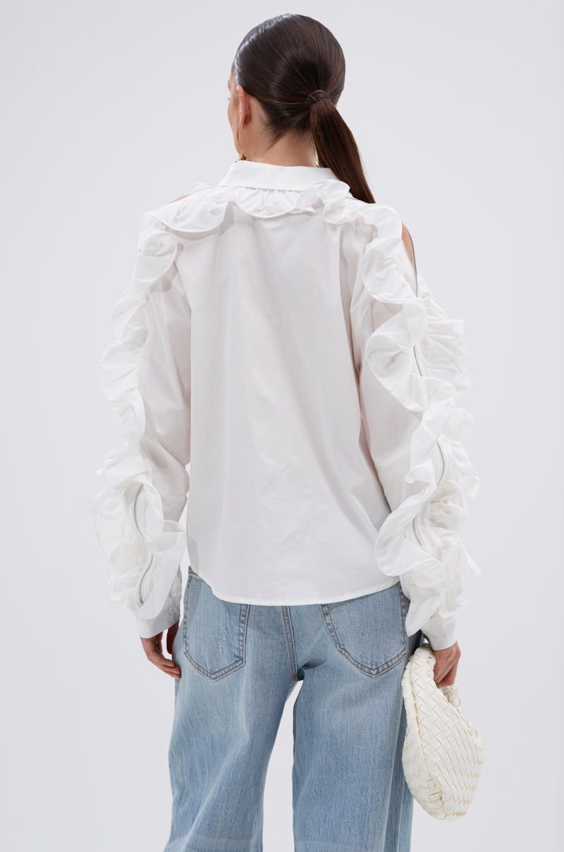 A woman with long dark hair in a low ponytail wears the ZIP ME OUT OF THIS BLOUSE with ruffle sleeve details and light blue jeans. She holds a white woven clutch, facing away to highlight the back against a plain background. 3