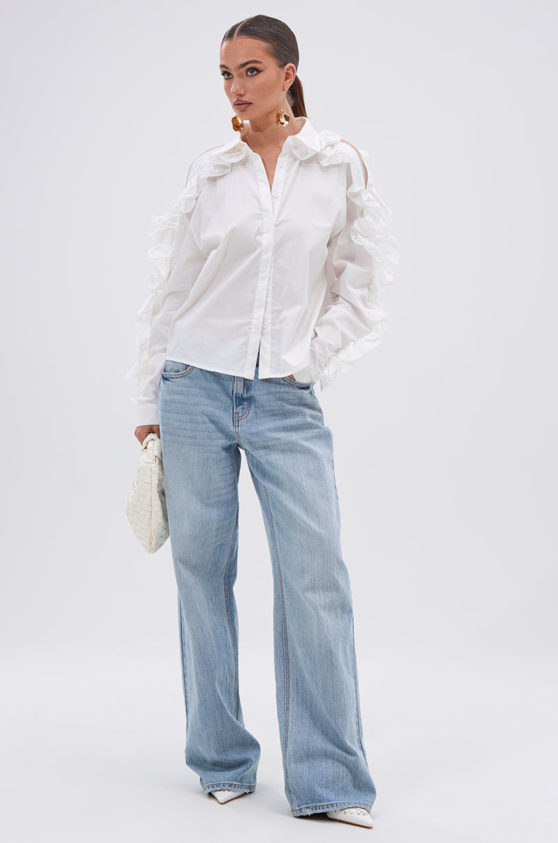 A woman with a sleek ponytail wears the ZIP ME OUT OF THIS BLOUSE with ruffled shoulders, paired with light blue wide-leg jeans, white pointed shoes, and a textured white handbag against a white background. 4