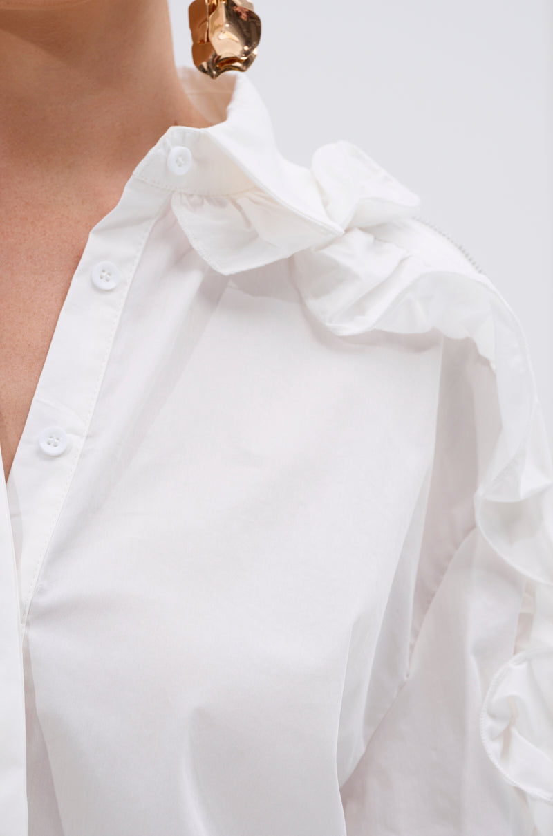 Close-up of a person in the ZIP ME OUT OF THIS BLOUSE, showcasing ruffle shoulder details and a metallic gold statement earring. The simple, light backdrop accentuates the blouse's sculptural design. 5