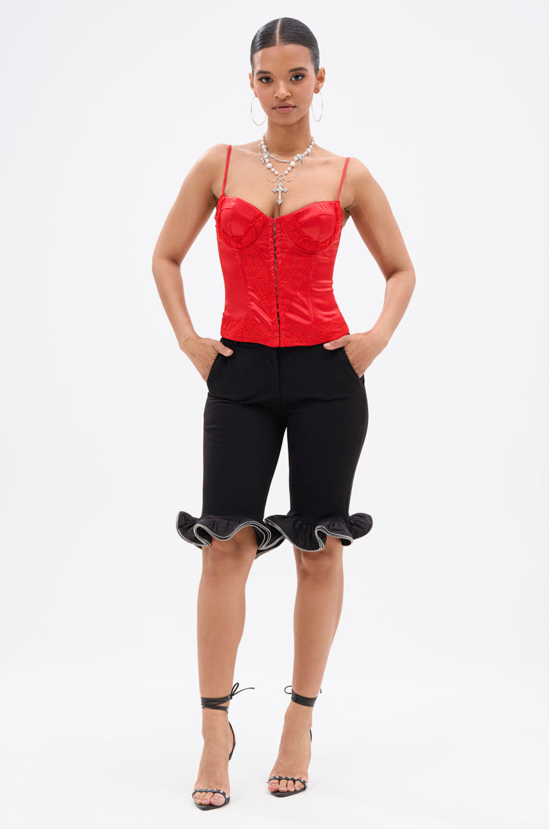 A woman stands confidently in the EXQUISITE SATIN LACE CORSET with structural boning, paired with black knee-length ruffled shorts, black strappy heels, and layered silver necklaces against a plain white background. 5