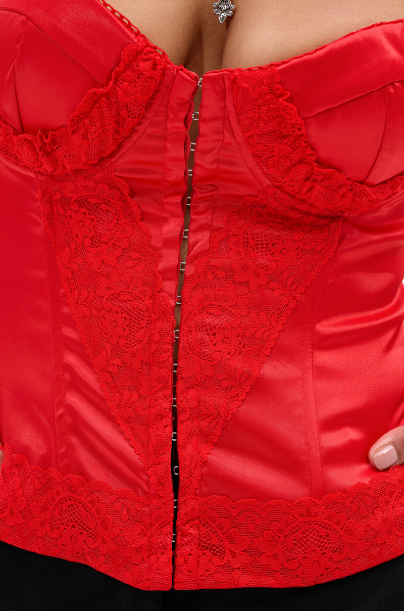 A close-up of someone wearing the EXQUISITE SATIN LACE CORSET in bright red, featuring structural boning, a front hook-and-eye closure, intricate lace patterns, and detailed stitching. A heart-shaped pendant necklace is partly visible at the top. 7