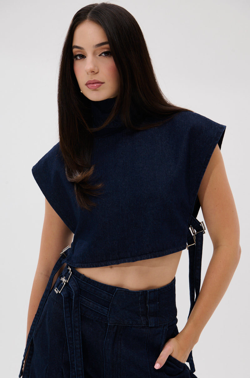A woman with long dark hair models the YOU A REAL ONE DENIM BLOUSE, a sleeveless top with side buckles, paired with matching high-waisted pants. She poses against a plain light background, one hand in her pocket.
