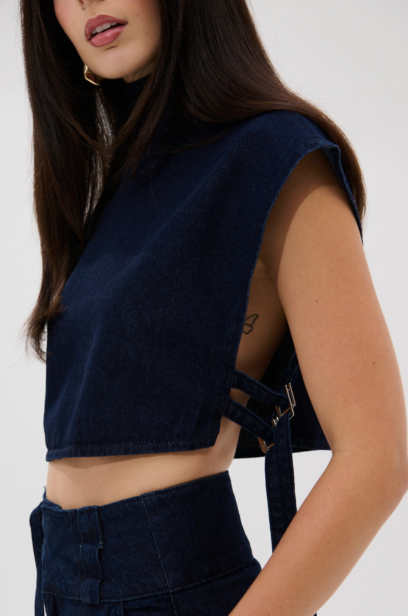 A woman wears the YOU A REAL ONE DENIM BLOUSE, a dark sleeveless top with buckle details, paired with matching high-waisted pants. Her long dark hair and a small tattoo under her arm are visible, though only part of her face shows.