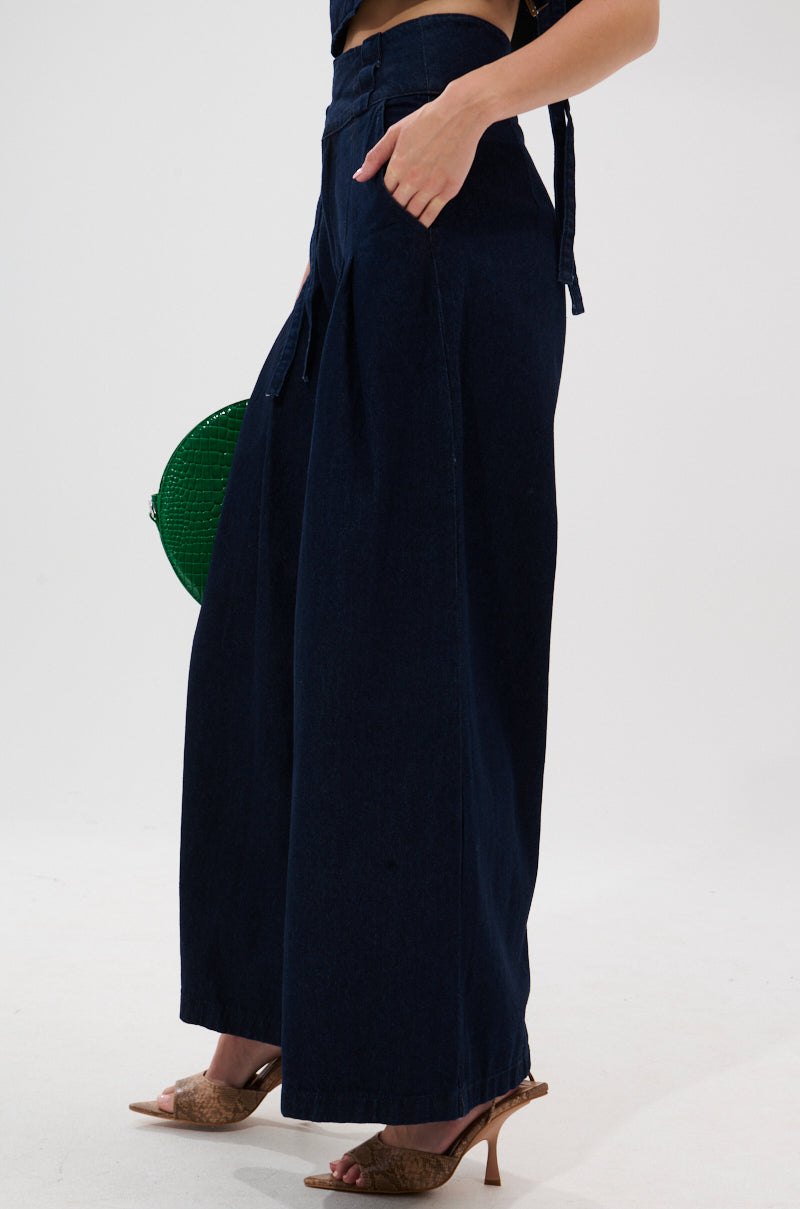 A person wearing the YOU A REAL ONE DENIM TROUSER stands sideways with hands in pockets, styled with brown open-toe heels, a round green bag, and a denim double buckle belt against a plain white background.