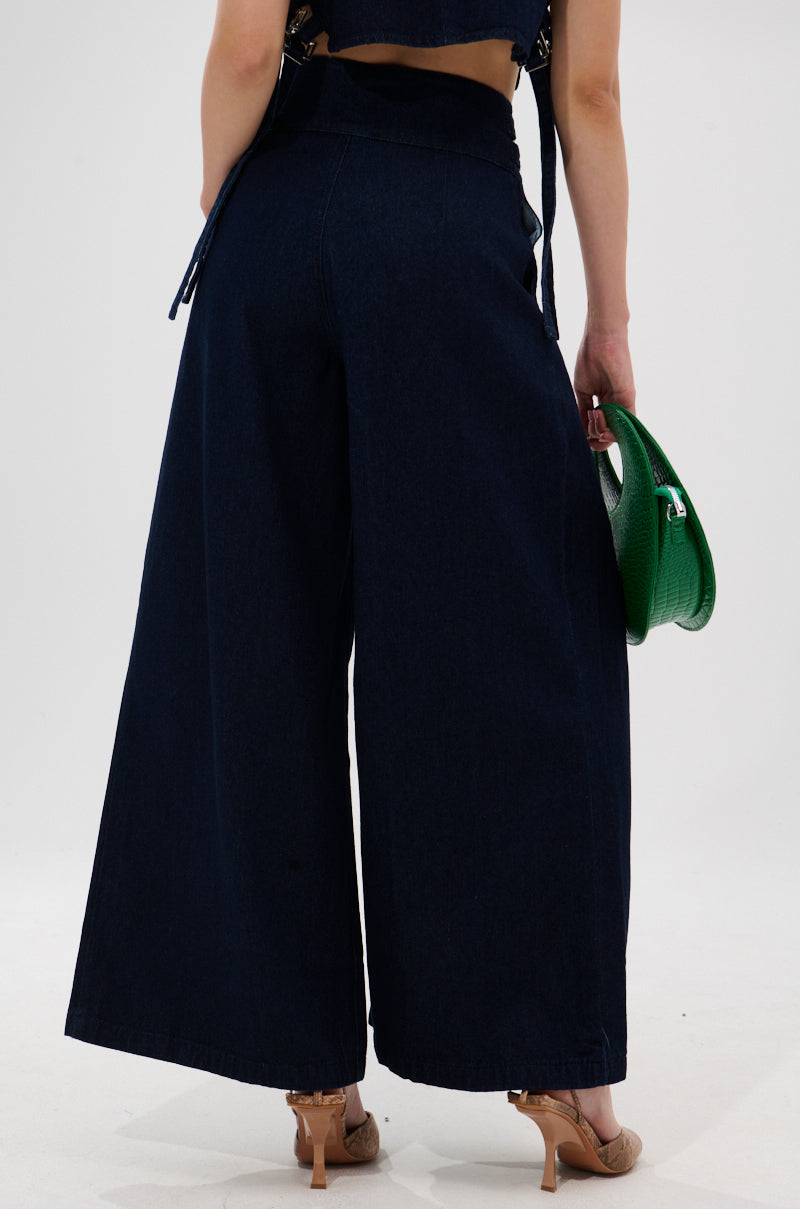 A person stands with their back to the camera, wearing YOU A REAL ONE DENIM TROUSER paired with tan heels and holding a small green handbag. The background is plain and light-colored.