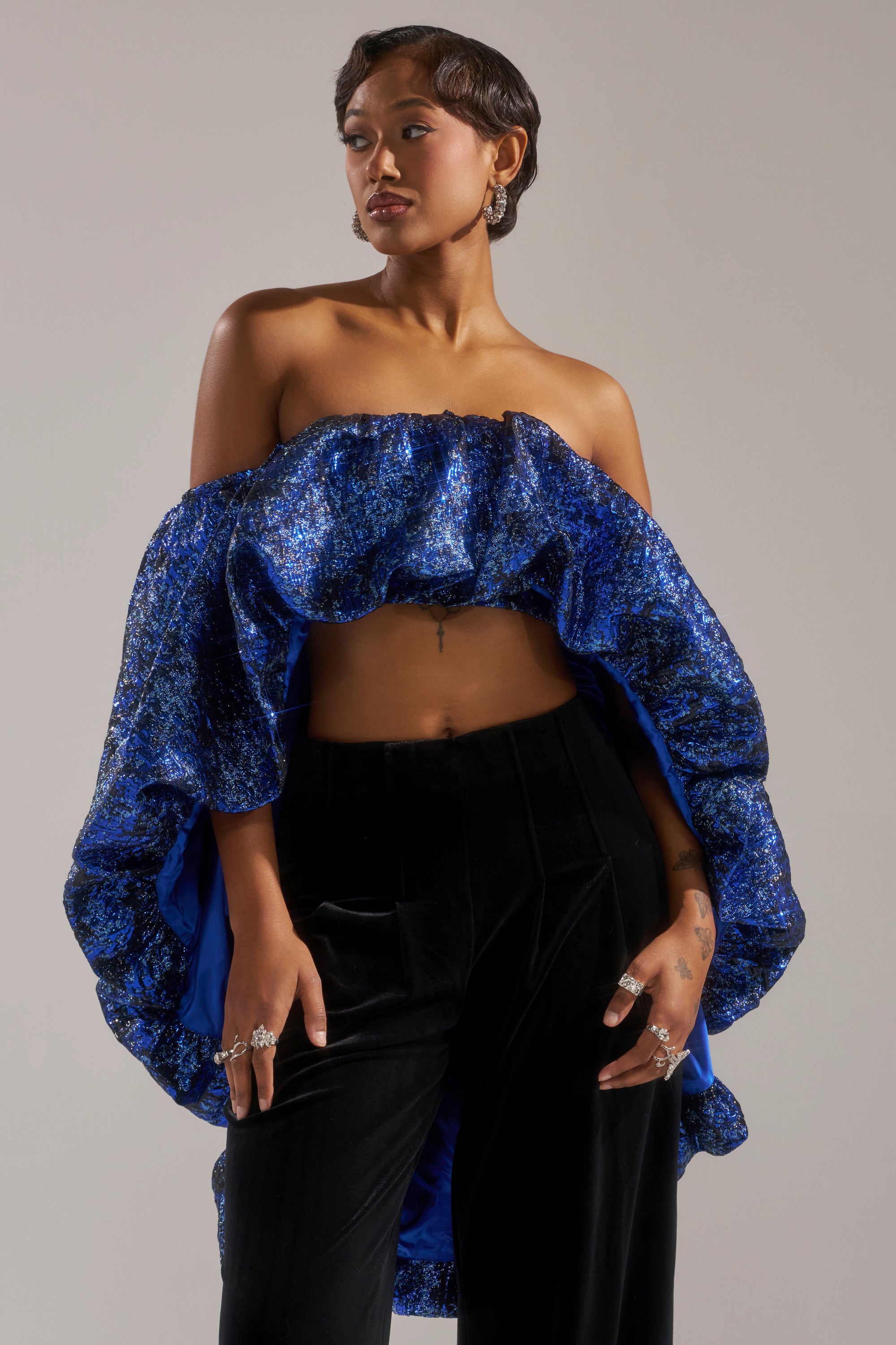 A woman with short, styled hair and a confident expression poses in the MELANI OFF THE SHOULDER HI-LOW BROCADE BLOUSE, paired with black velvet pants and silver rings and earrings against a neutral background.