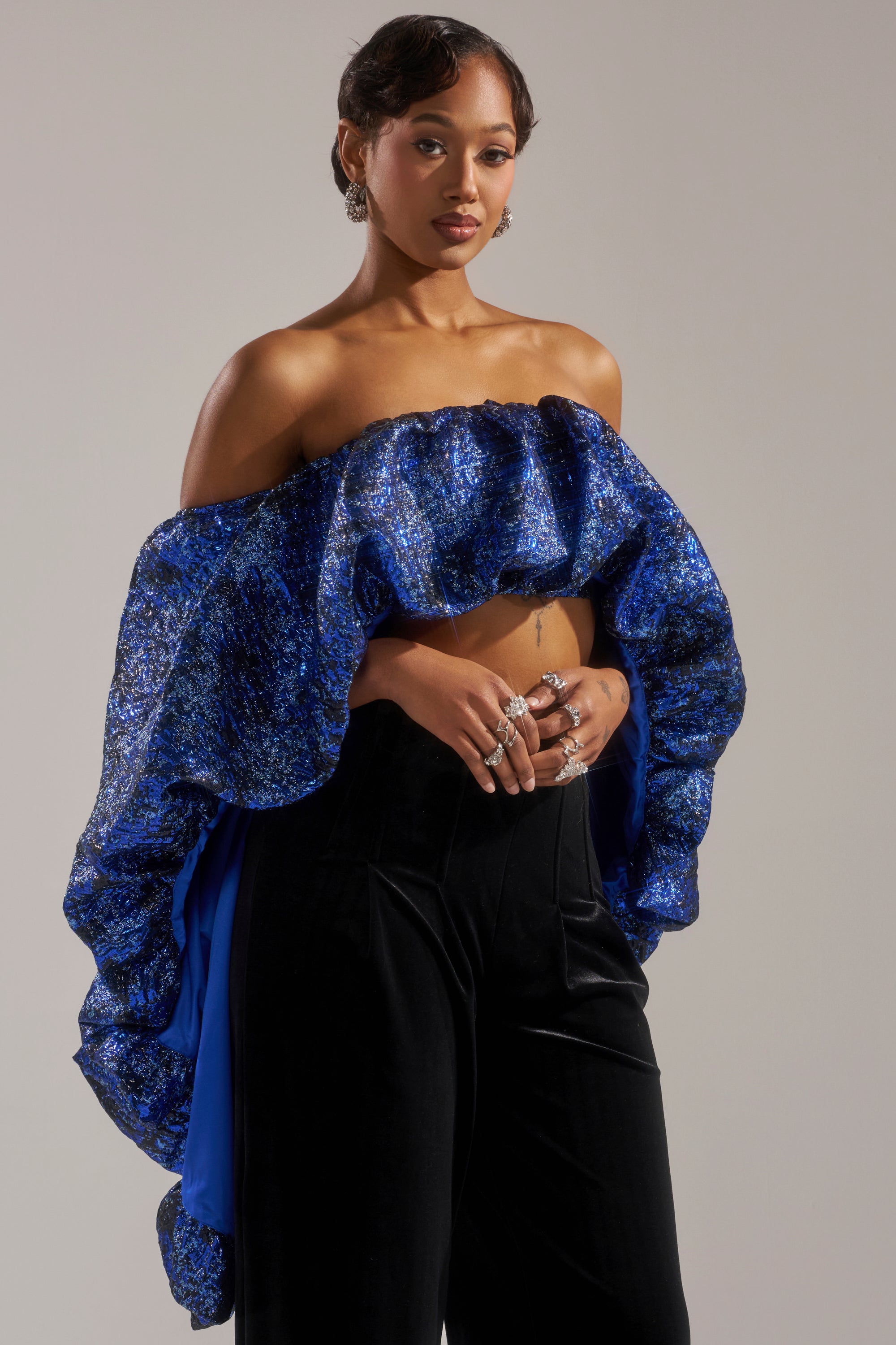 A woman poses against a plain background in the MELANI OFF THE SHOULDER HI-LOW BROCADE BLOUSE, featuring sparkly blue jacquard and ruffled details, paired with black velvet pants. She accessorizes with hoop earrings and rings.