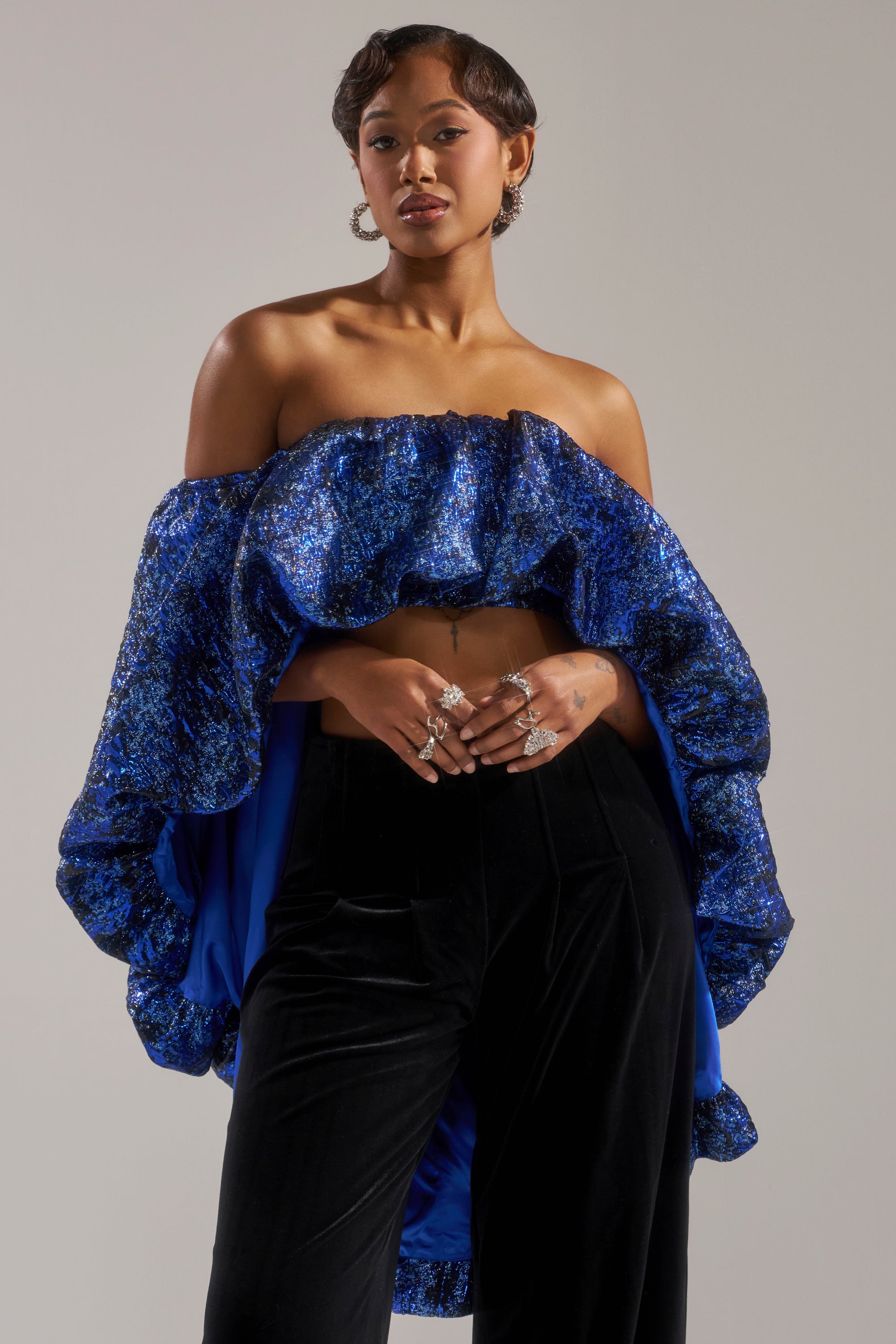 A woman with short hair, hoop earrings, and rings poses confidently in the MELANI OFF THE SHOULDER HI-LOW BROCADE BLOUSE, featuring a blue metallic shimmer, paired with black velvet pants against a neutral background.