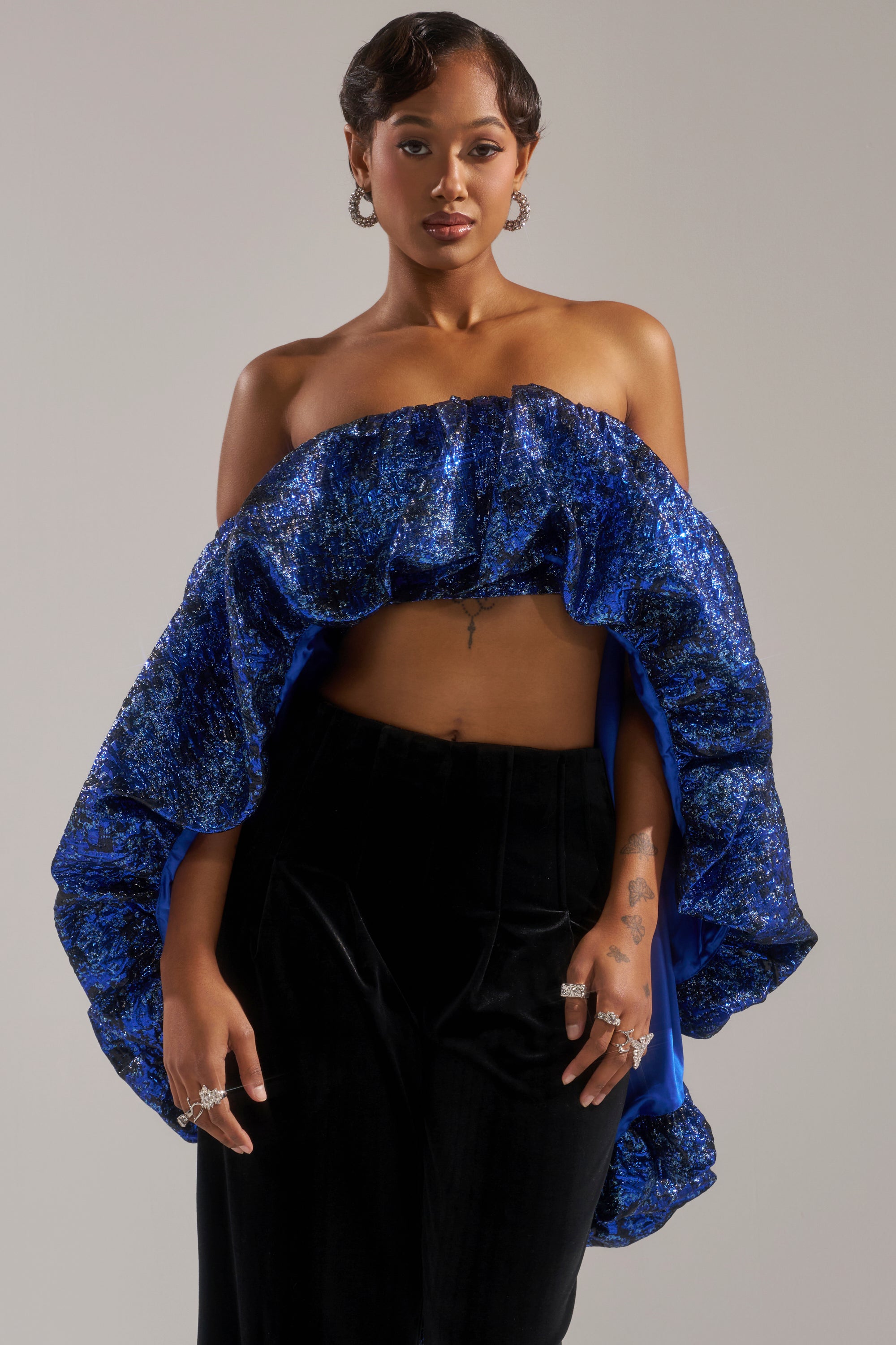 A woman poses confidently in the MELANI Off the Shoulder Hi-Low Brocade Blouse with royal blue ruffles, paired with high-waisted black pants. She sports short styled hair, hoop earrings, and several rings on her fingers.