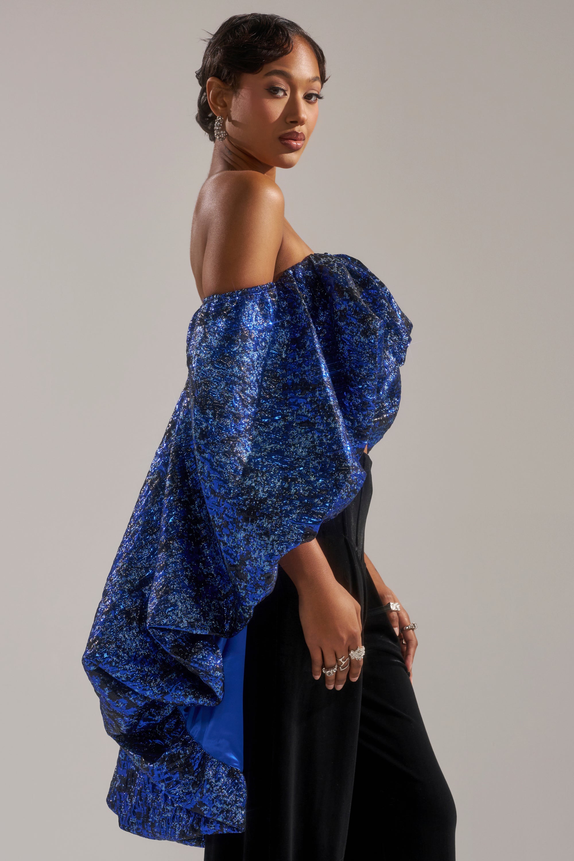 A woman poses in profile against a plain background, wearing the MELANI OFF THE SHOULDER HI-LOW BROCADE BLOUSE with blue textured ruffles and voluminous sleeves, paired with black pants. She has short hair and several rings.