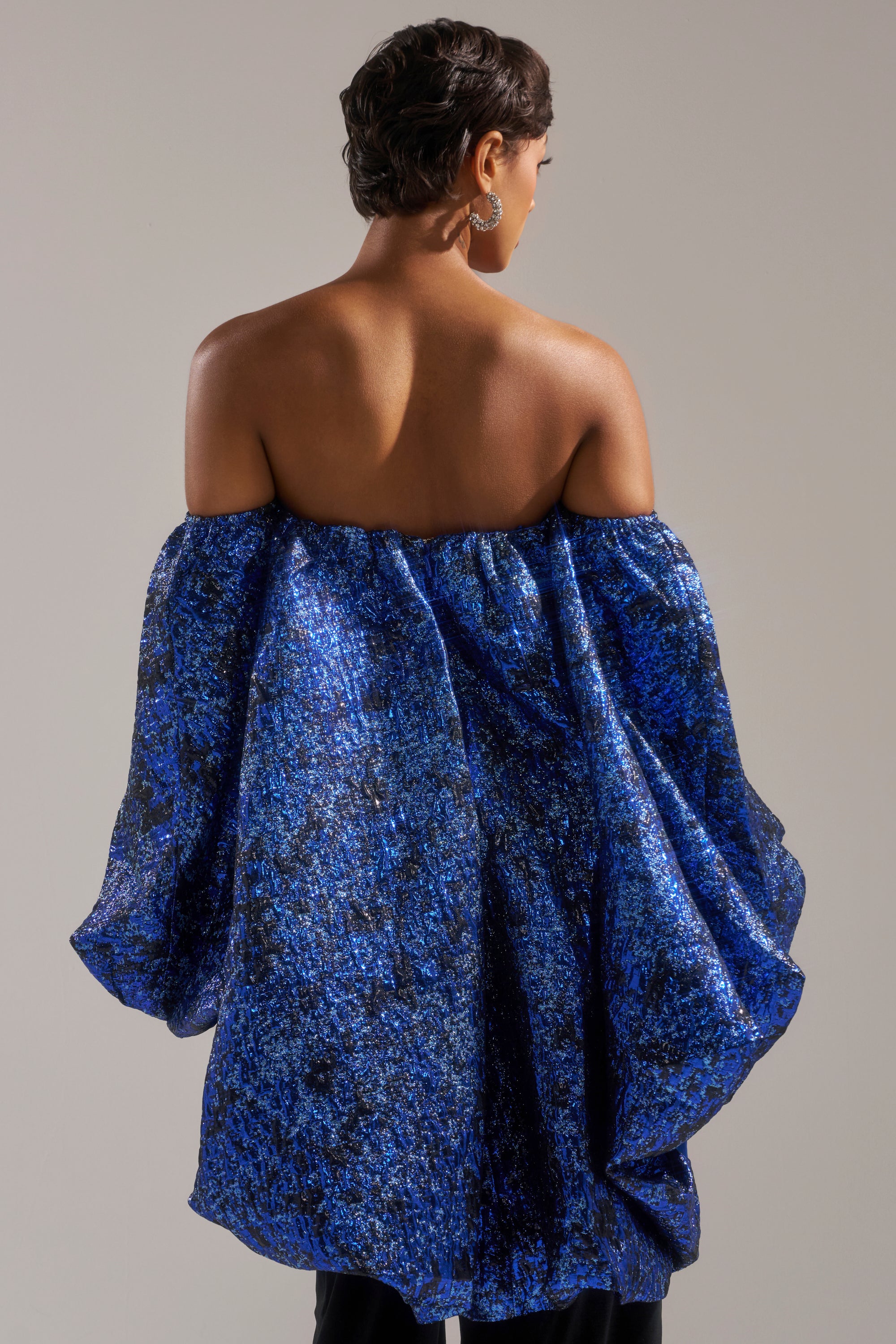 A person with short, dark hair is seen from behind wearing the MELANI OFF THE SHOULDER HI-LOW BROCADE BLOUSE in textured blue and black against a plain, neutral background.