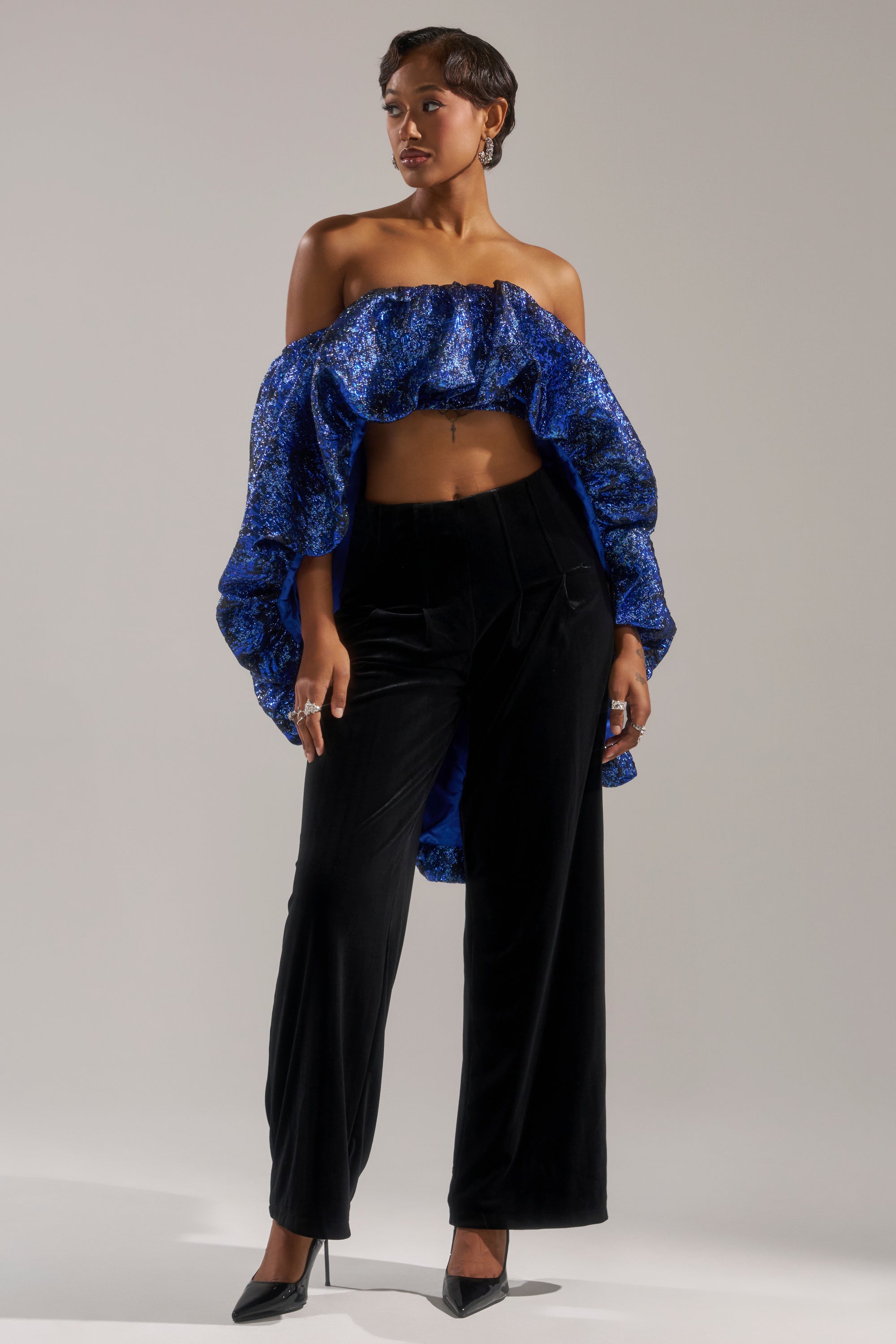 A person poses in the MELANI OFF THE SHOULDER HI-LOW BROCADE BLOUSE, featuring dramatic ruffled sleeves, paired with high-waisted black velvet pants, black pointed heels, short hair, rings, and earrings.