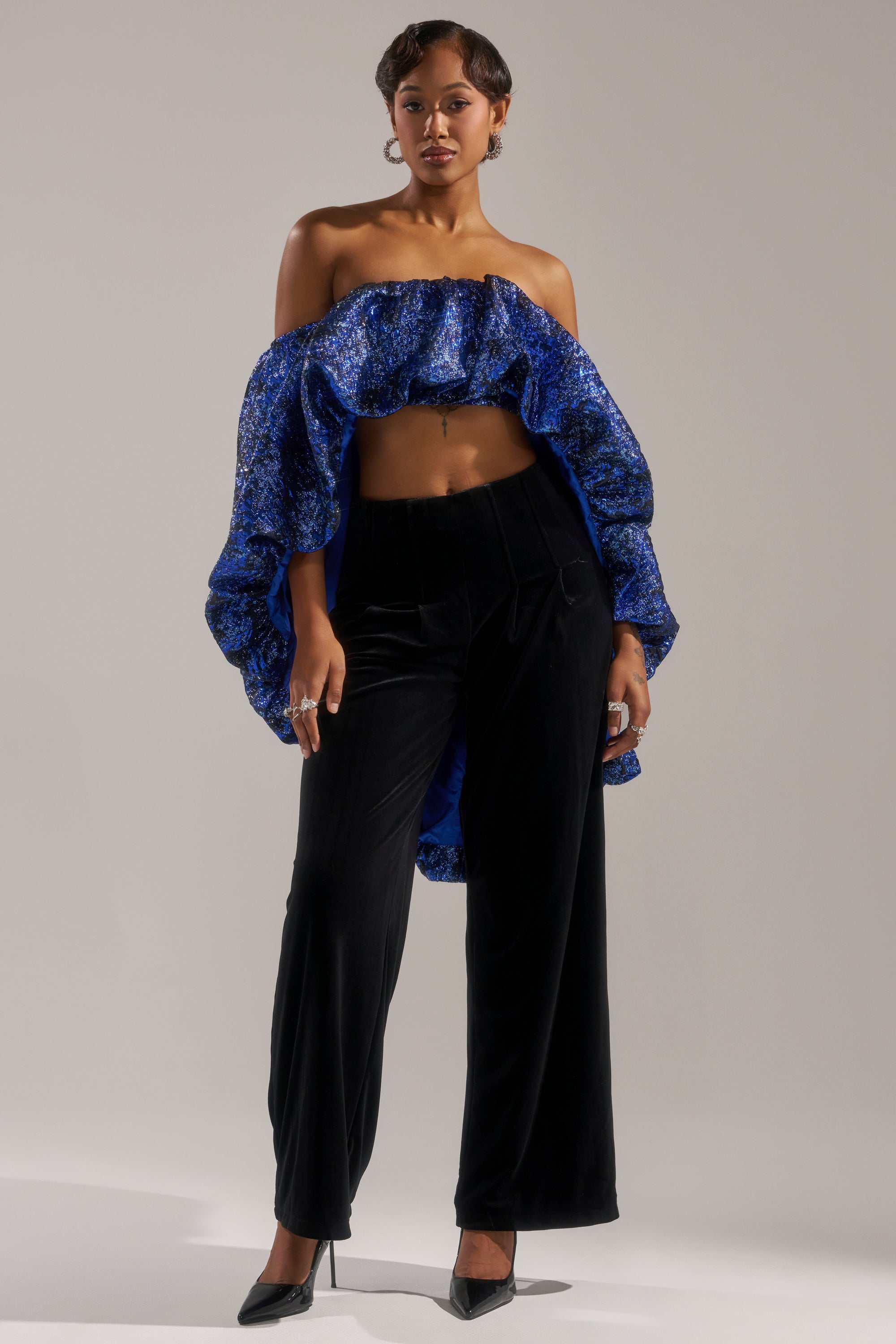 A woman exudes confidence in the MELANI OFF THE SHOULDER HI-LOW BROCADE BLOUSE, styled with high-waisted black velvet pants and black pointed heels. The striking blue hue stands out against the neutral background.