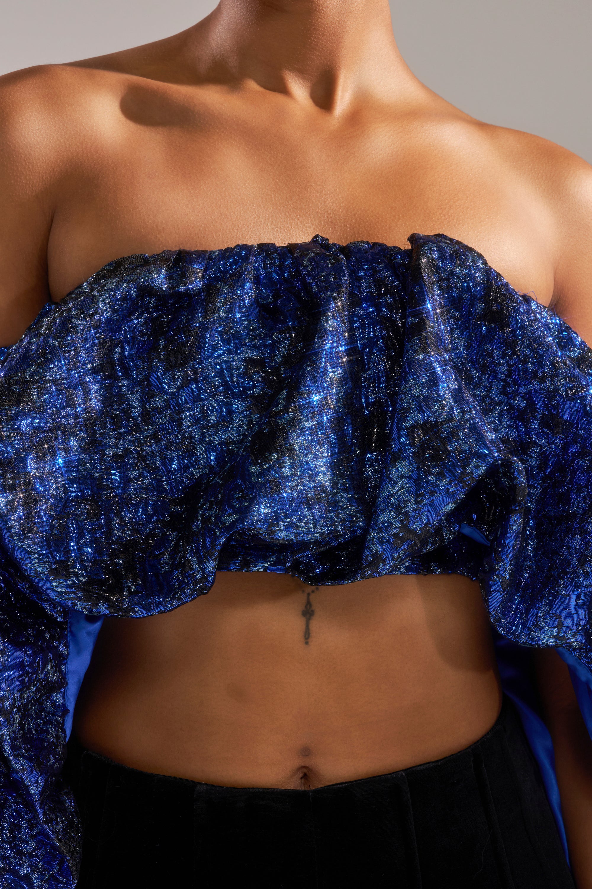 A person wears the MELANI OFF THE SHOULDER HI-LOW BROCADE BLOUSE in blue and black jacquard, showcasing bare shoulders and arms, with a small cross tattoo visible on the upper abdomen below the chest.