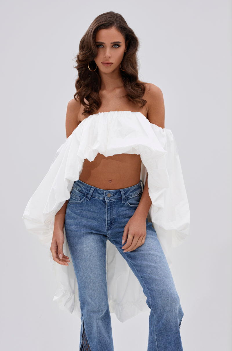 A woman with wavy brown hair and large hoop earrings wears the MELANI OFF THE SHOULDER HIGH LOW BLOUSE in lightweight fabric with a draped back, paired with blue jeans, against a plain light background. 0
