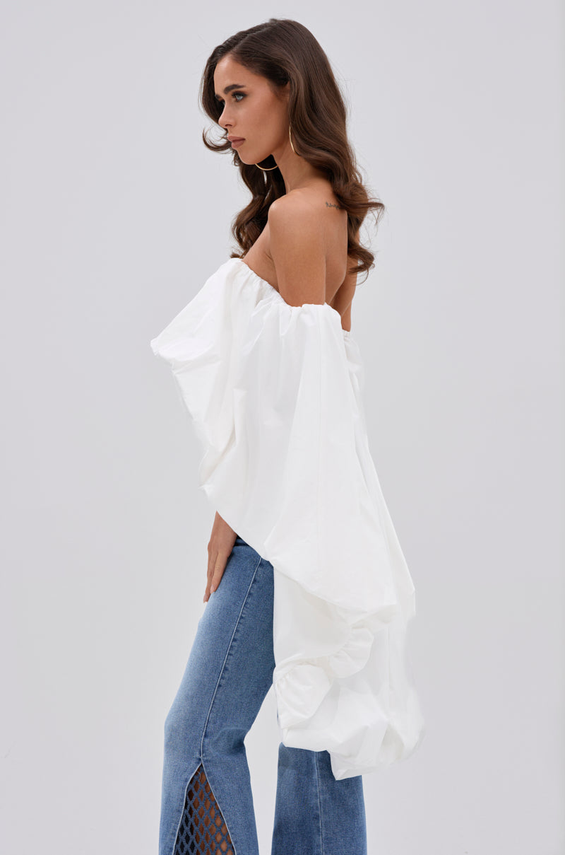 A woman stands sideways in the MELANI OFF THE SHOULDER HIGH LOW BLOUSE, a lightweight, flowy long-sleeve top, paired with blue jeans with split-hem detail. The background is plain and light-colored. 2
