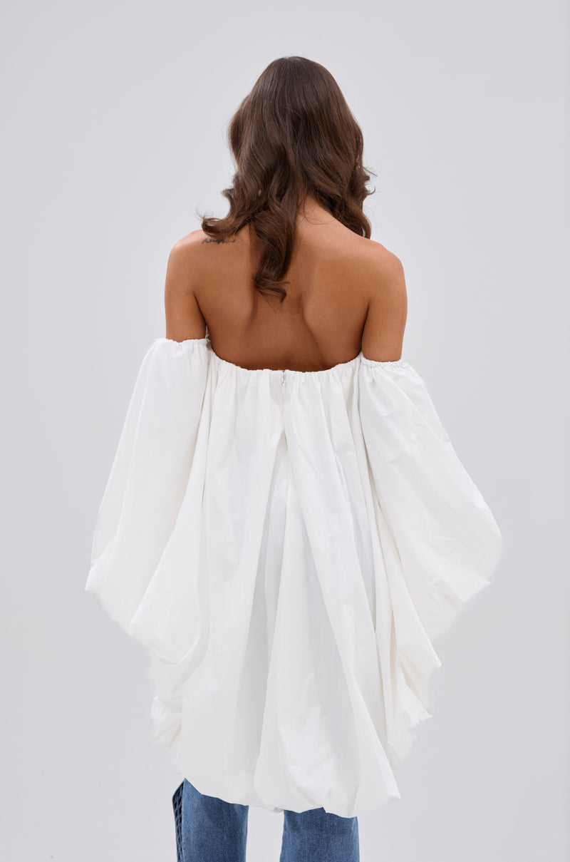 A woman with long, wavy brown hair stands facing away in the MELANI OFF THE SHOULDER HIGH LOW BLOUSE, made from lightweight fabric and paired with blue jeans against a plain light gray background. 3