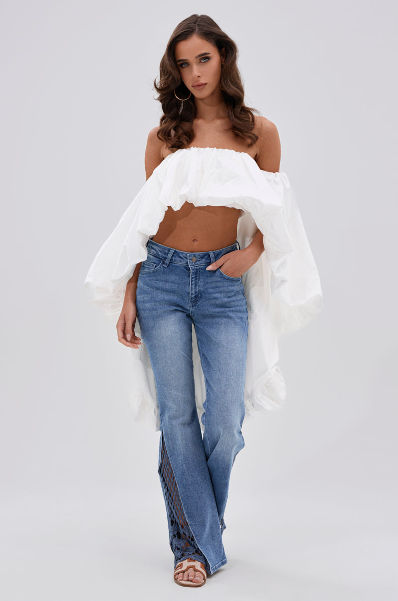 A woman with long wavy hair wears the MELANI OFF THE SHOULDER HIGH LOW BLOUSE and blue jeans with ankle cutouts, standing against a plain light gray background with one hand in her pocket. 4