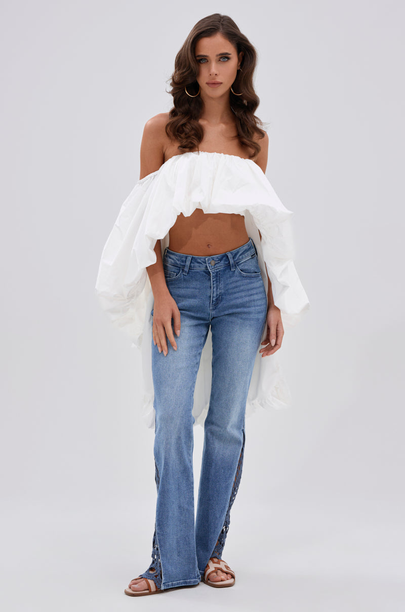 A woman with wavy brown hair wears the MELANI OFF THE SHOULDER HIGH LOW BLOUSE paired with blue jeans and brown sandals. The lightweight white top with ruffled details stands out against a light gray background. 6