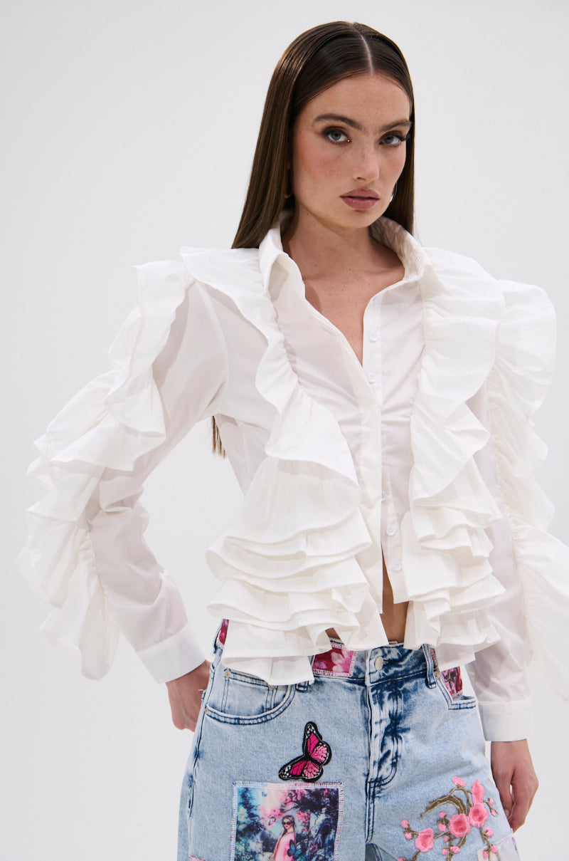 A woman with straight brown hair models the STAR OF THE SHOW RUFFLE BUTTON DOWN, a white ruffle shirt, paired with light blue jeans adorned with colorful patches like a pink butterfly and floral designs, against a plain white background.