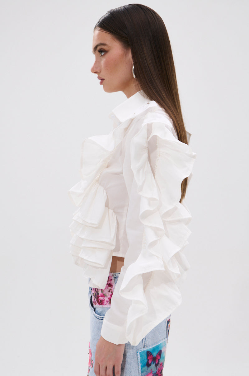 A woman with straight brown hair models the STAR OF THE SHOW RUFFLE BUTTON DOWN, paired with blue jeans featuring colorful floral patches, standing in profile against a plain white background.