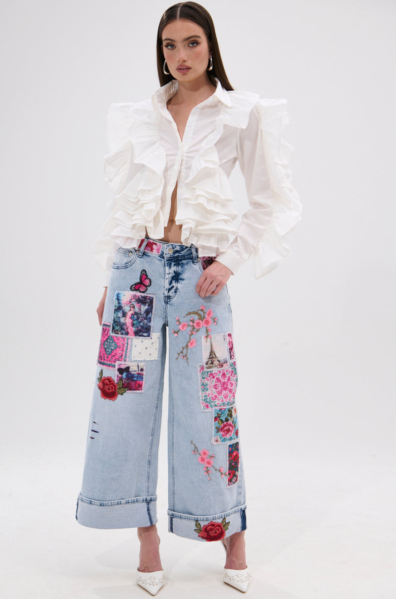 A woman stands against a white background wearing the STAR OF THE SHOW RUFFLE BUTTON DOWN and wide-leg light blue jeans with colorful patches, embroidery, and floral designs, completed with white pointed heels for a statement look.