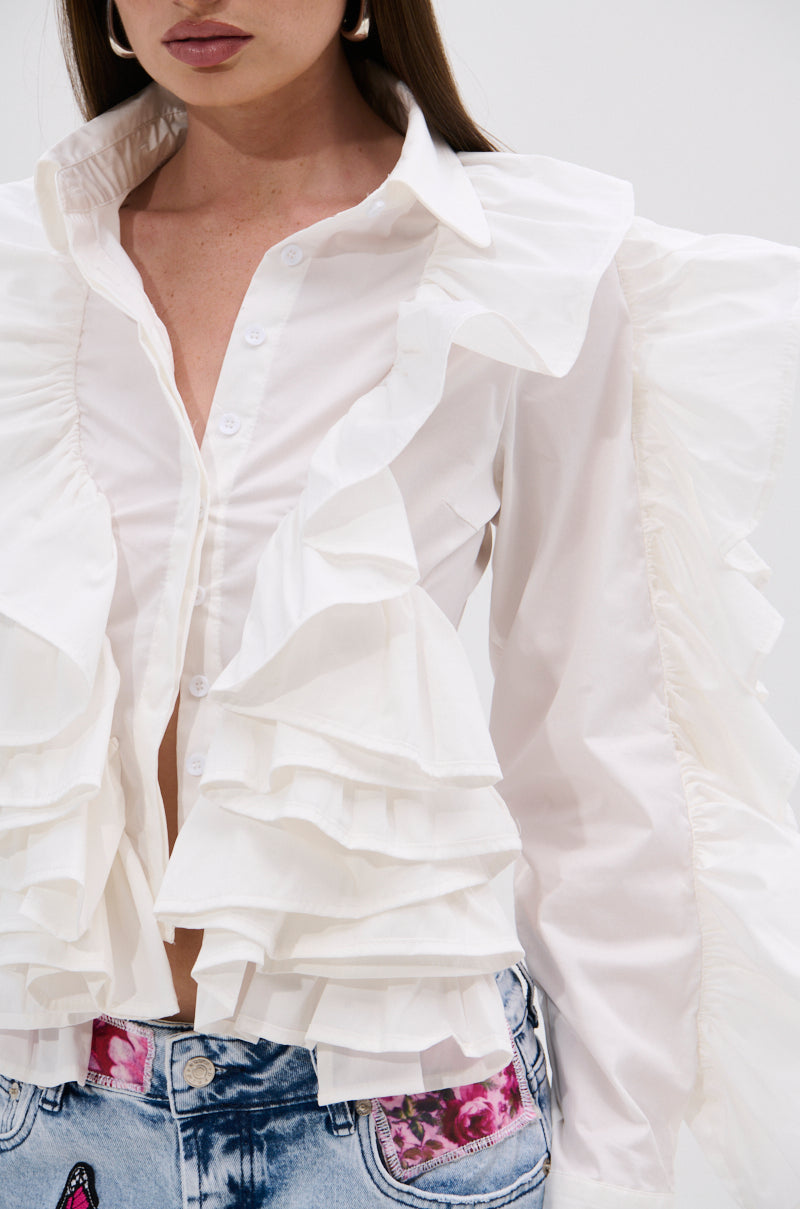 A woman models the STAR OF THE SHOW RUFFLE BUTTON DOWN, a white blouse with layered ruffles, paired with light blue jeans adorned with pink floral patches. The cropped image captures her from shoulders to hips.