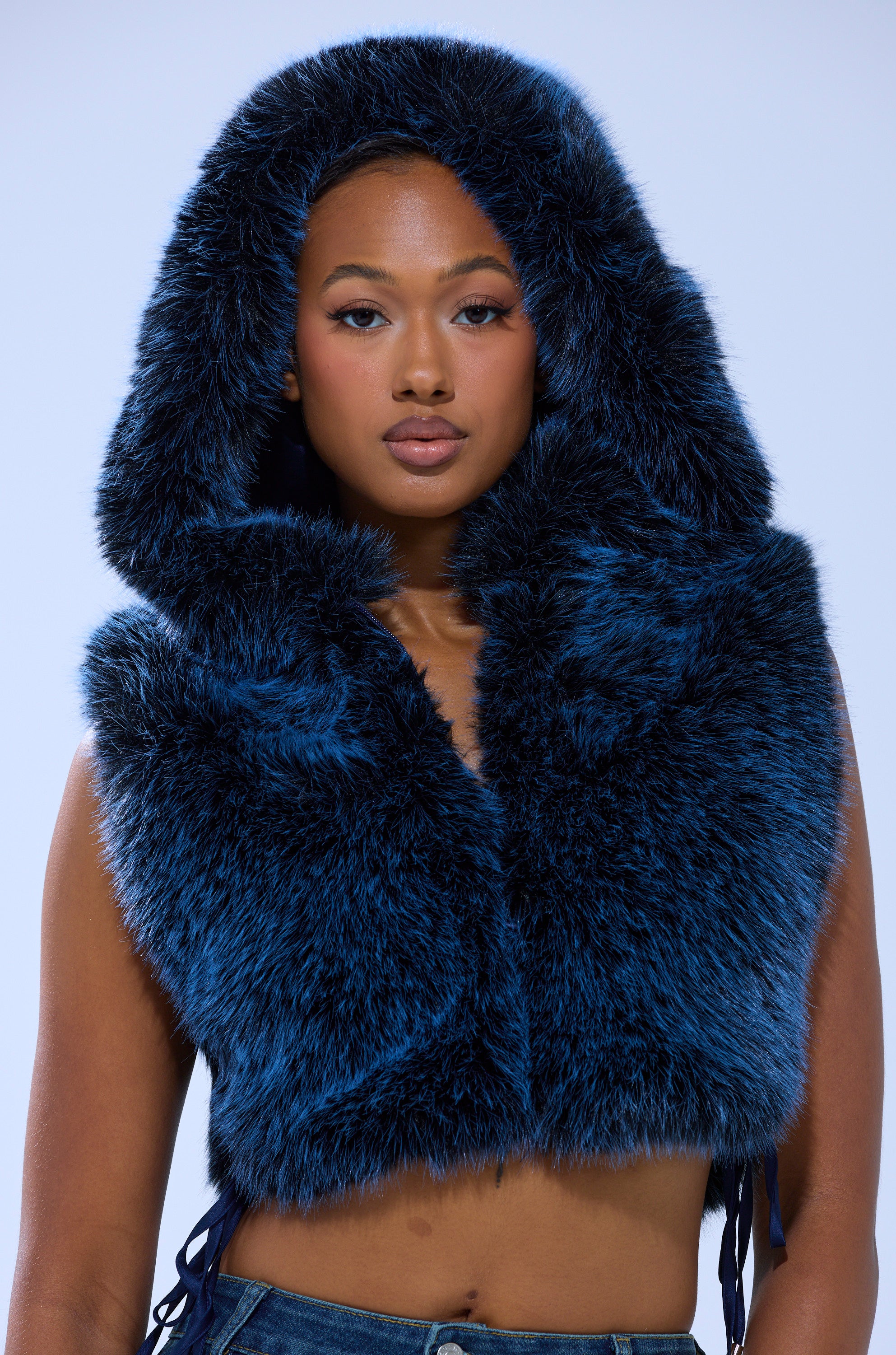 A woman wearing the ALL A DREAM FUR TOP— a sleeveless, hooded, dark blue faux fur piece inspired by Y2K fashion—stands against a plain light background, gazing at the camera with a neutral expression.