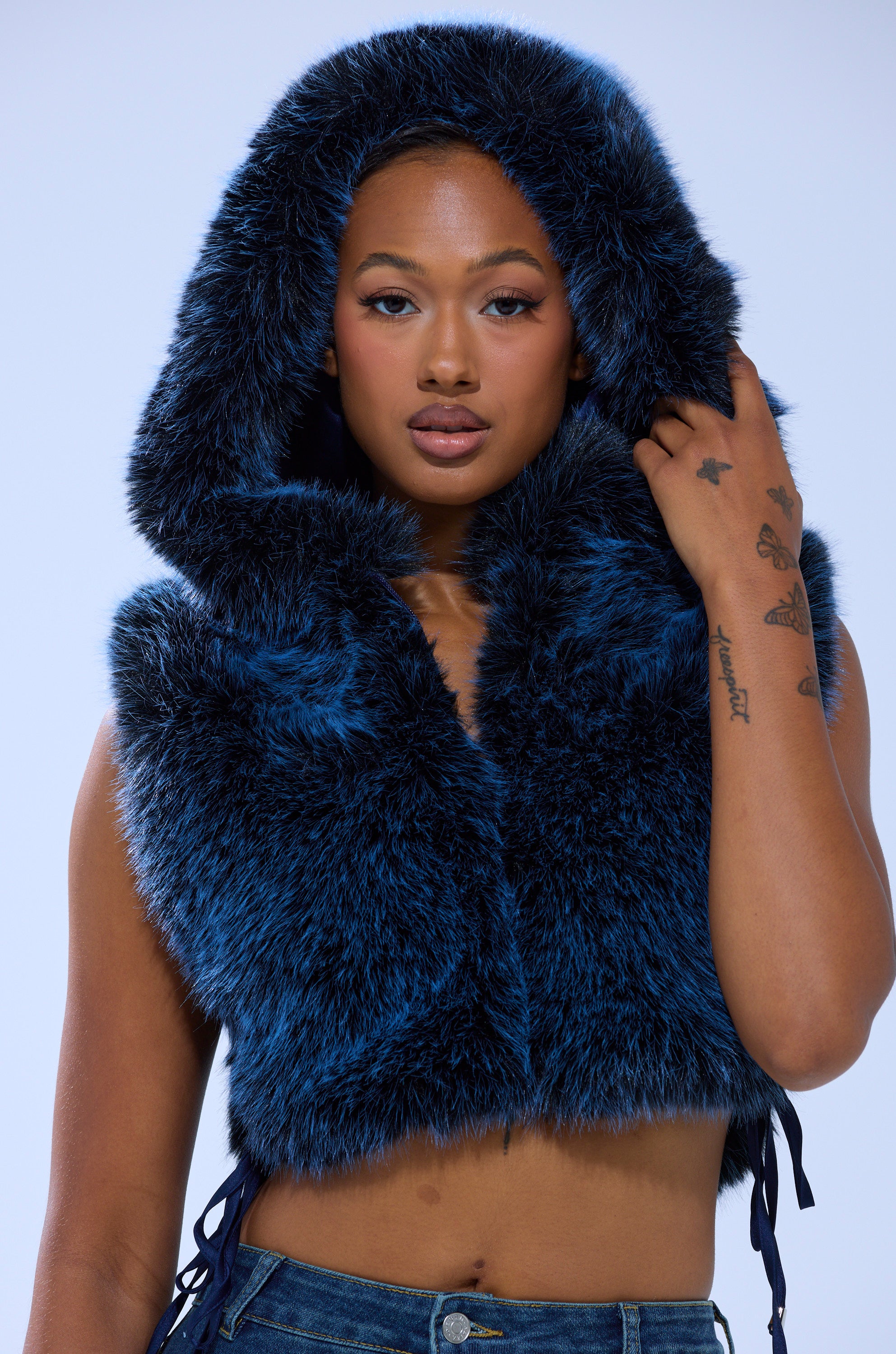 A woman in the blue ALL A DREAM FUR TOP by AKIRA Label, featuring a sleeveless design and hood, looks at the camera while holding the hood’s edge and showing butterfly tattoos on her hand. The background is plain and light-colored.
