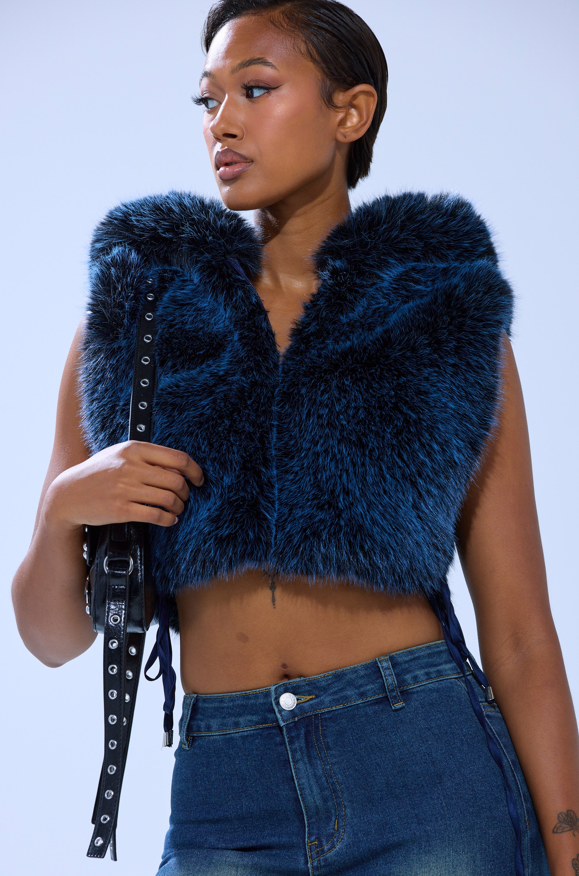 A woman wears the sleeveless blue ALL A DREAM FUR TOP with blue jeans, holding a black strap over her shoulder against a light blue background.