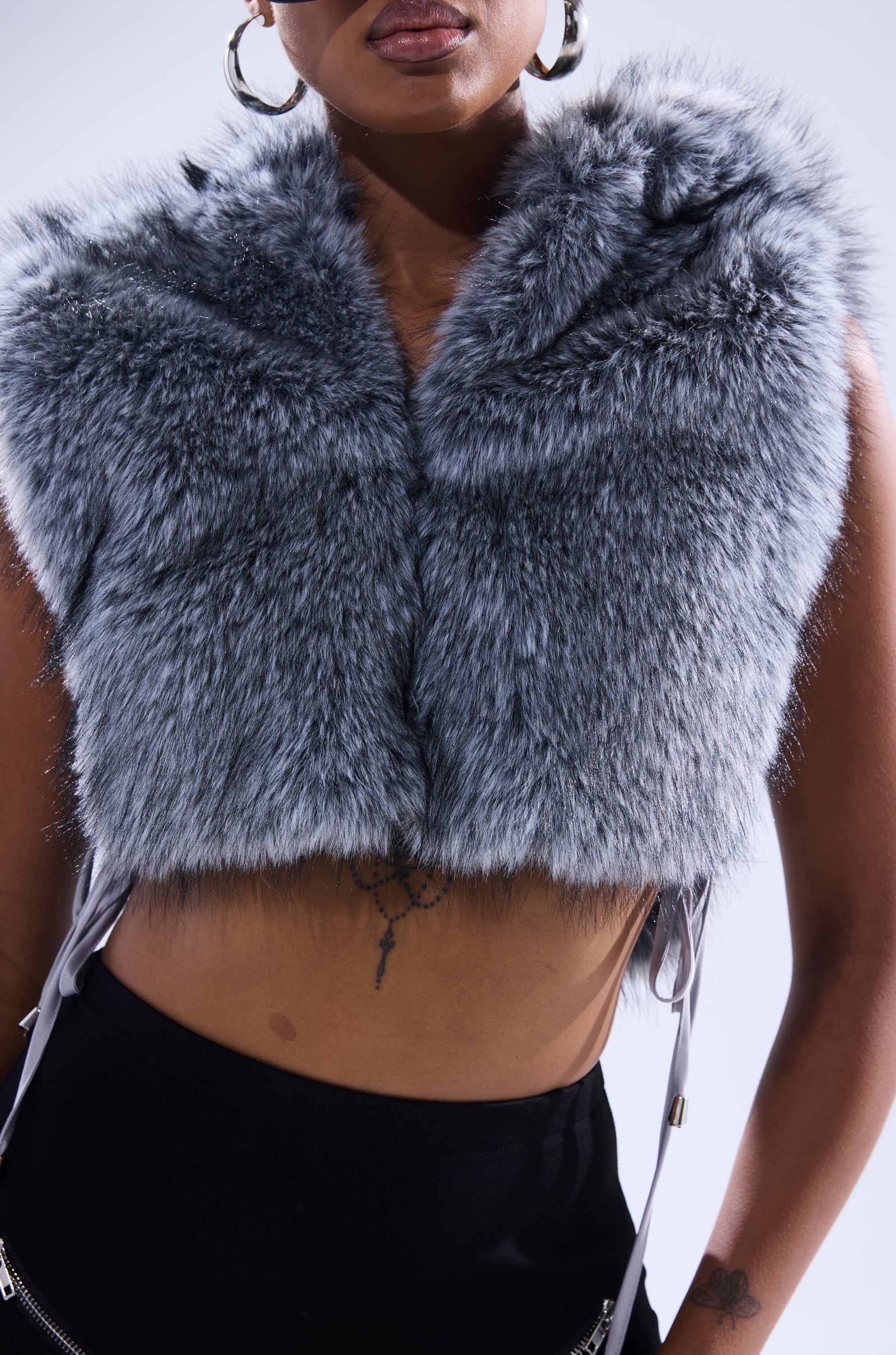 A person models the TOUCH ME FUR TOP—sleeveless, gray, and hooded—paired with black pants and large hoop earrings, revealing a tattoo on their midriff against a plain background. 9