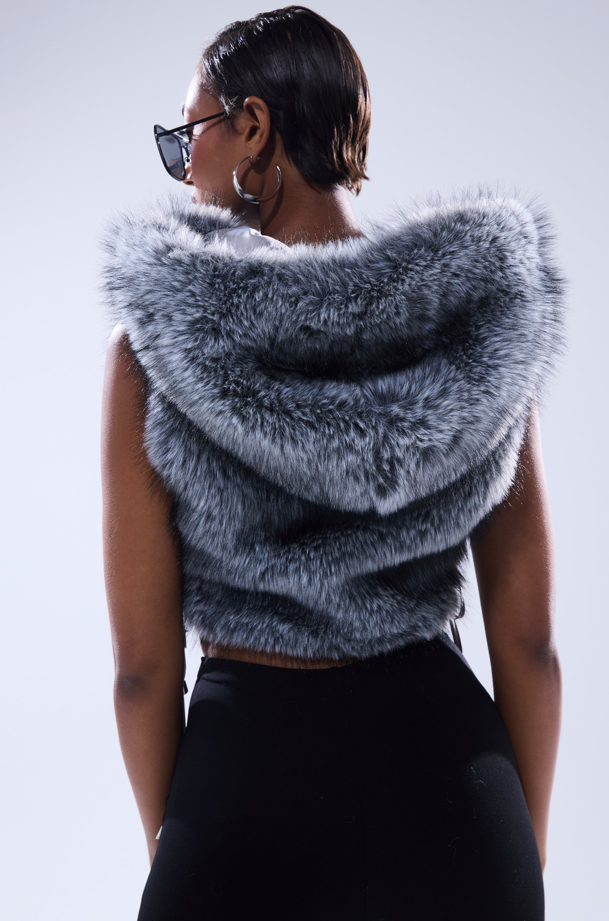 A woman with short hair and hoop earrings faces away from the camera in sunglasses and the TOUCH ME FUR TOP, a sleeveless hooded faux fur top, set against a plain light background. 6