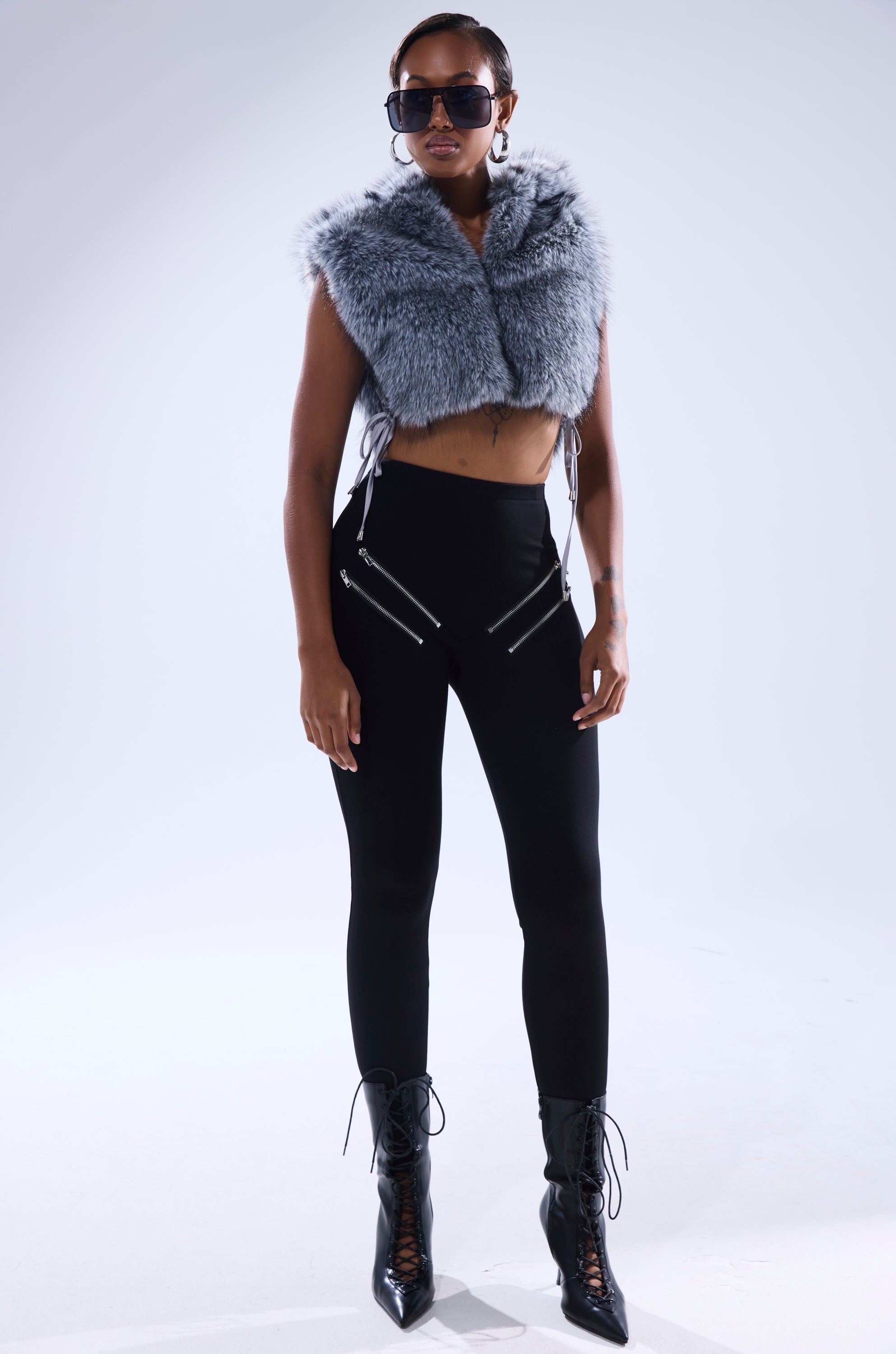 A woman stands confidently against a plain light background, wearing sunglasses, the TOUCH ME FUR TOP styled as a gray cropped vest, high-waisted black zipper-detailed pants, and black lace-up ankle boots. 7